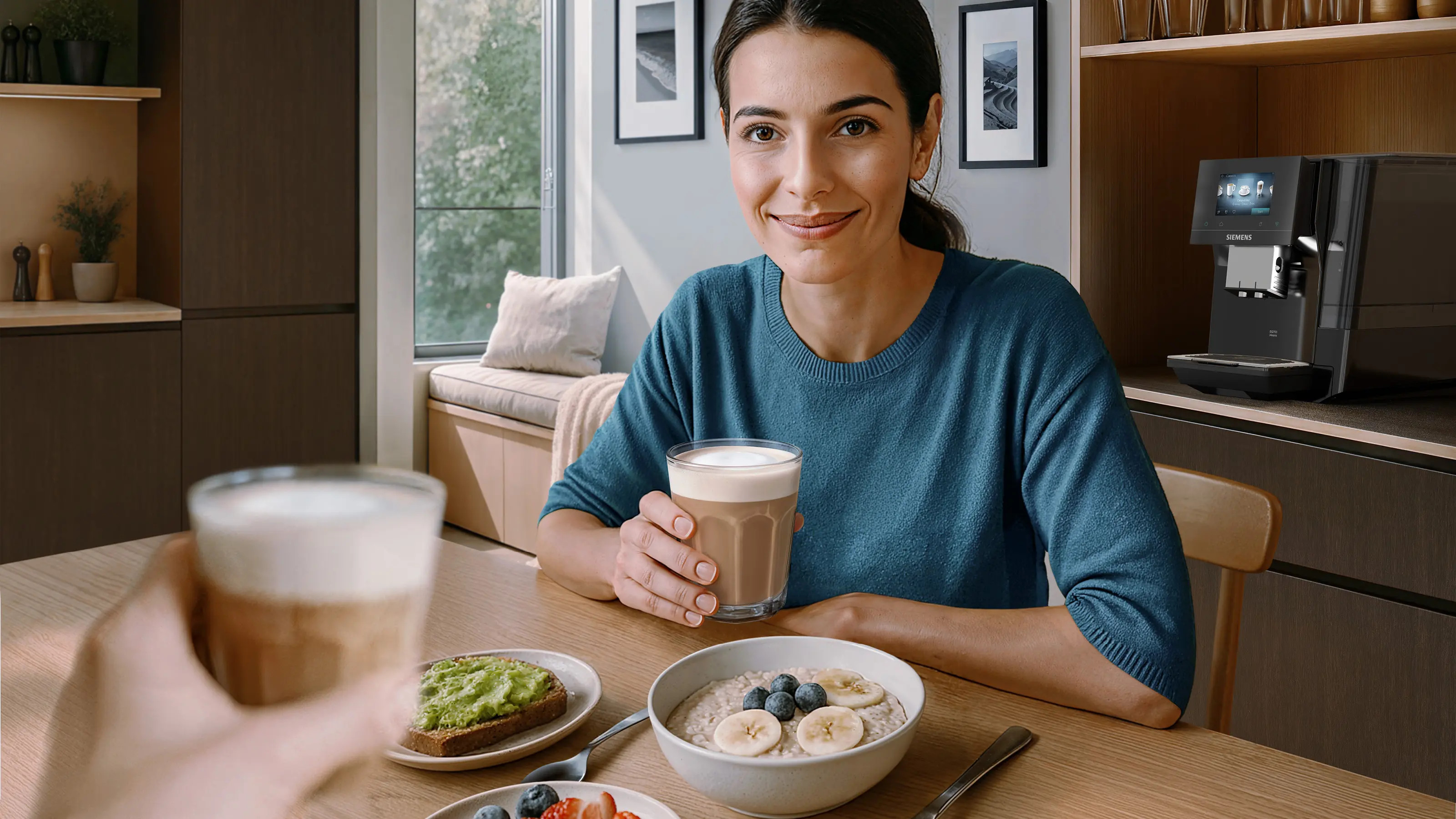 Kobieta z kawą i śniadaniem przy stole z ekspresem do kawy w tle.