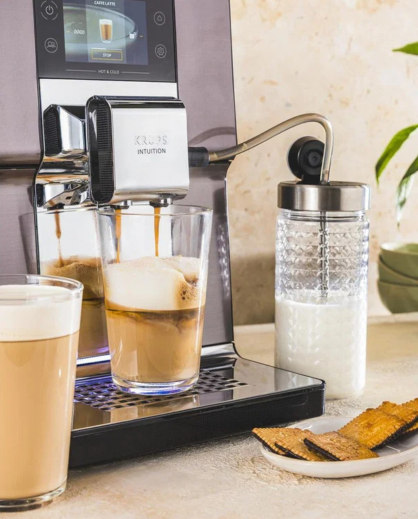 Ekspres do kawy parzy kawę do szklanki, obok dzbanek na mleko i ciastka.