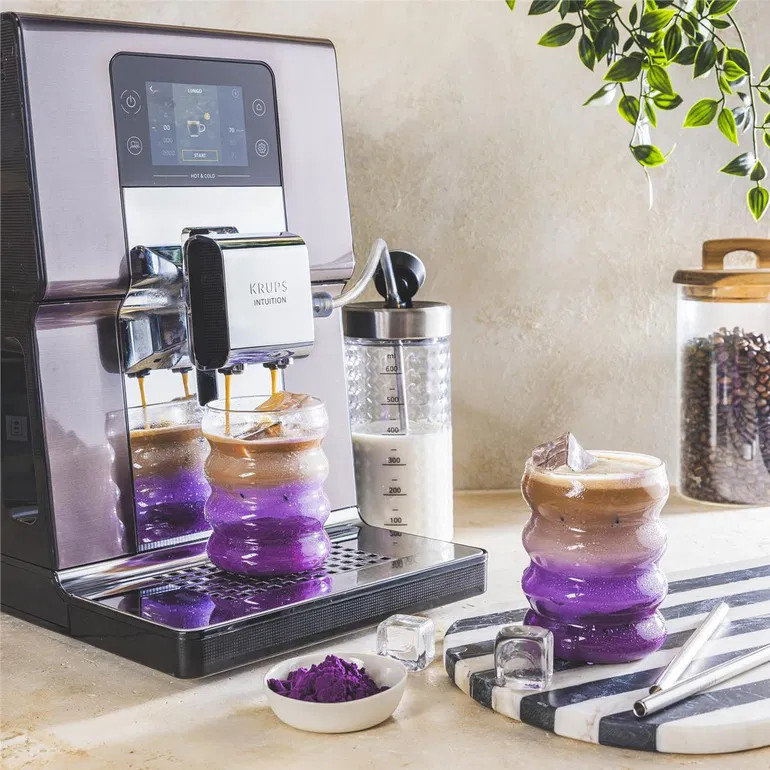 Ekspres do kawy przygotowuje dwukolorowy napój w szklance. Ziarna kawy w tle.