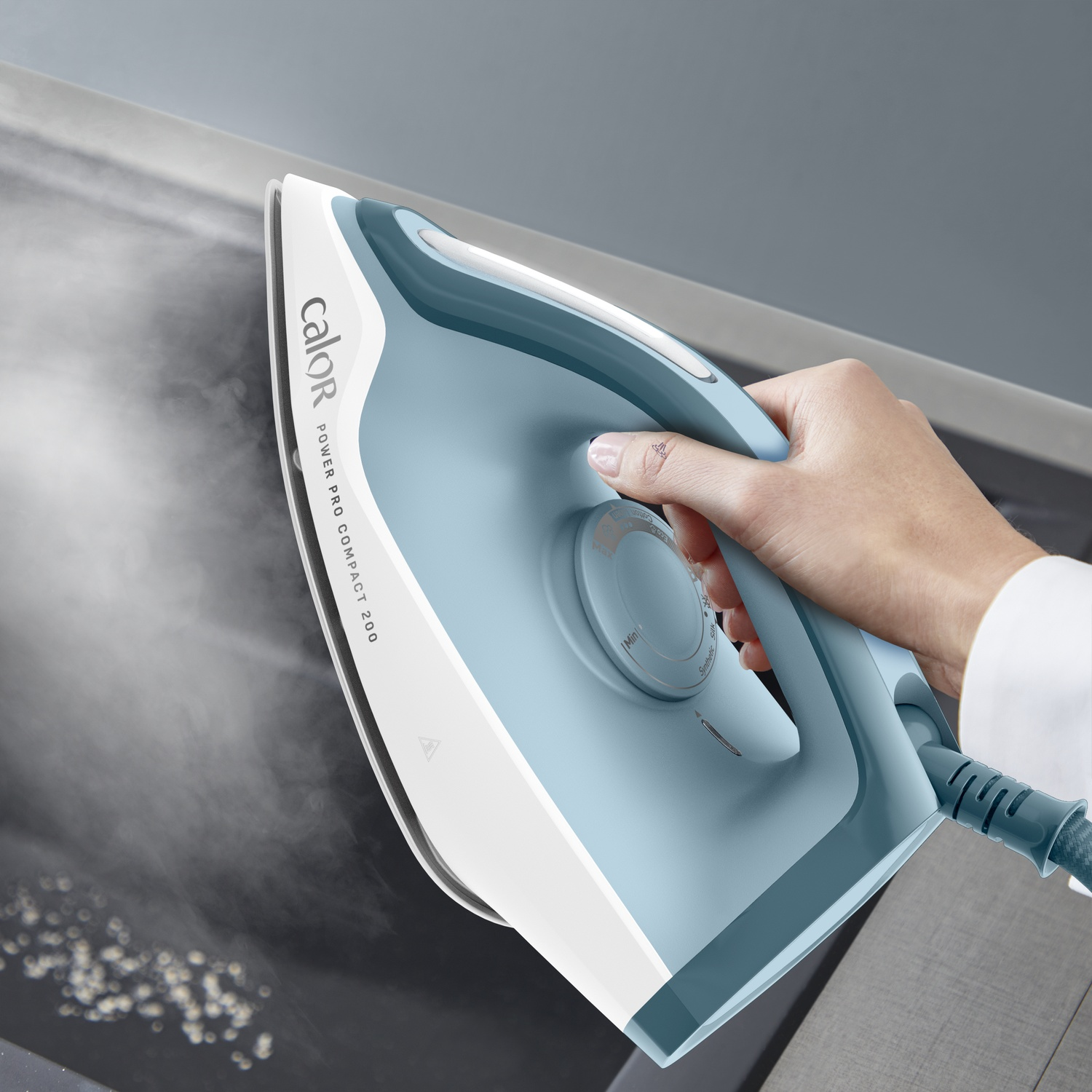 A hand holds a blue and white iron, with steam emitting from the soleplate.