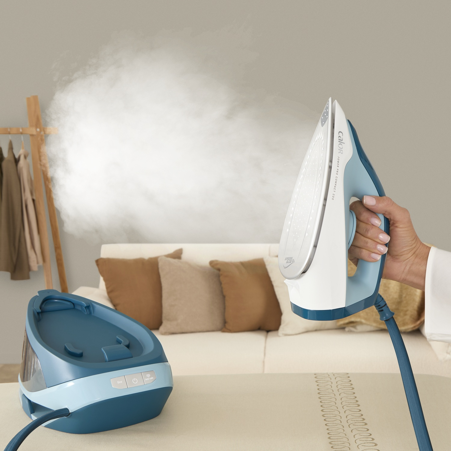 A hand holds an iron releasing steam. An iron stand is in the background.