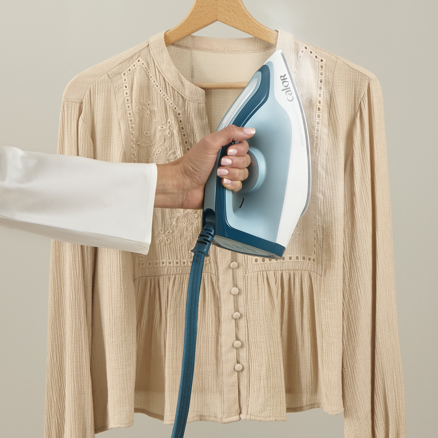 A hand irons a beige blouse with steam, hanging on a hanger.