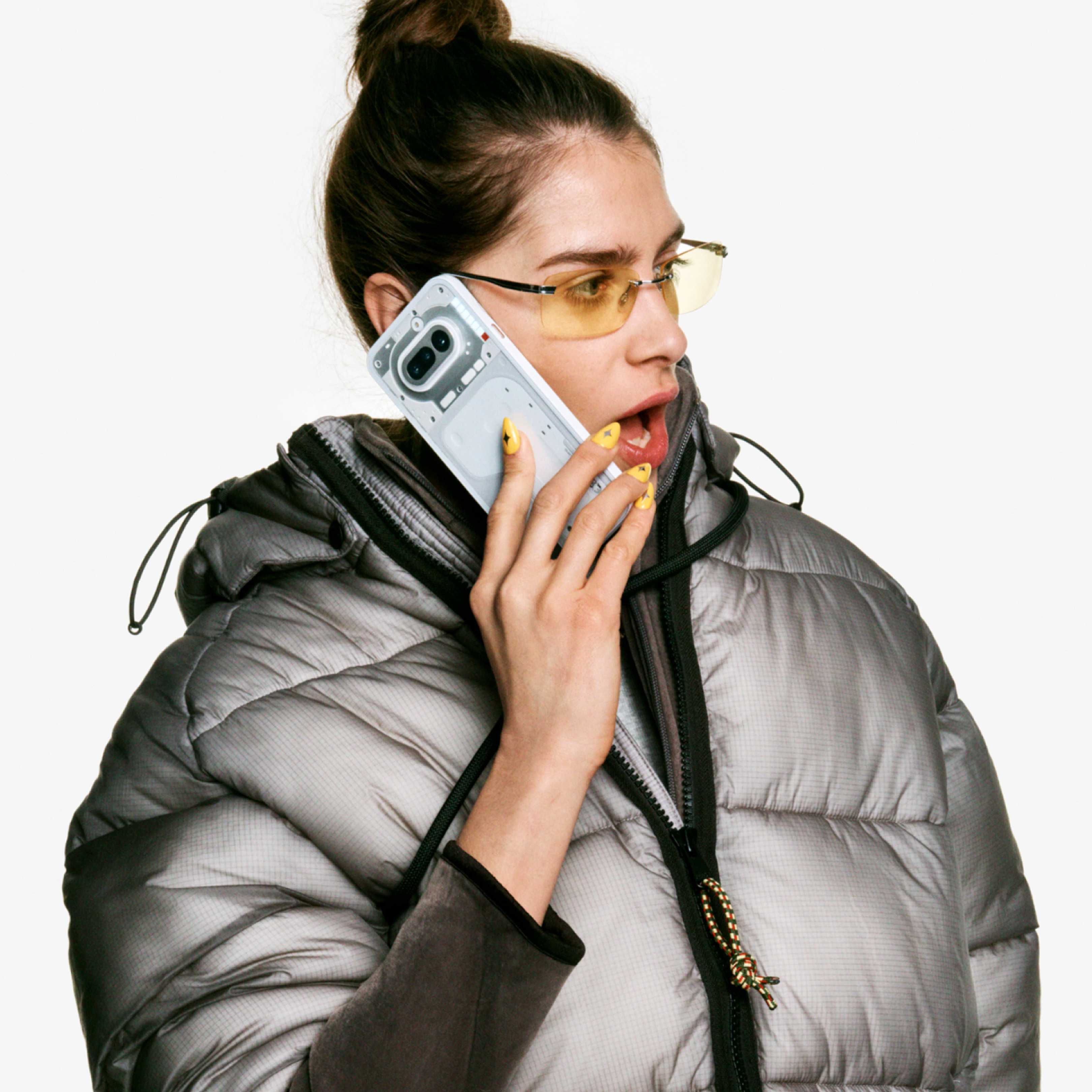 Frau mit gelben Brillen telefoniert mit einem transparenten Handy, trägt eine Pufferjacke.