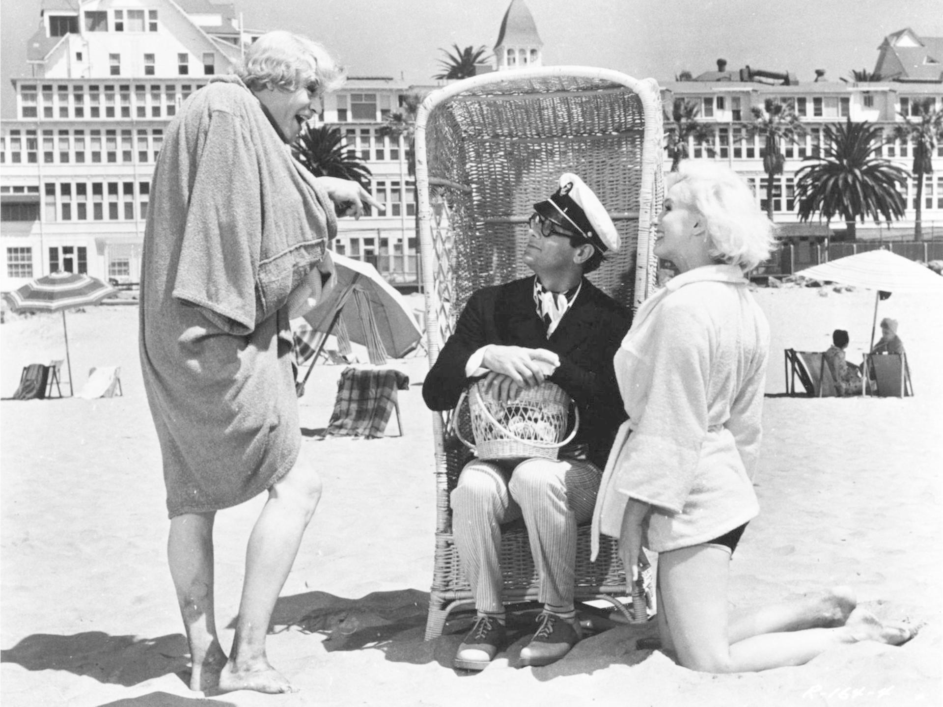 Drei Personen am Strand vor einem Gebäude mit Palmen. Einer sitzt in einem Korbsessel.