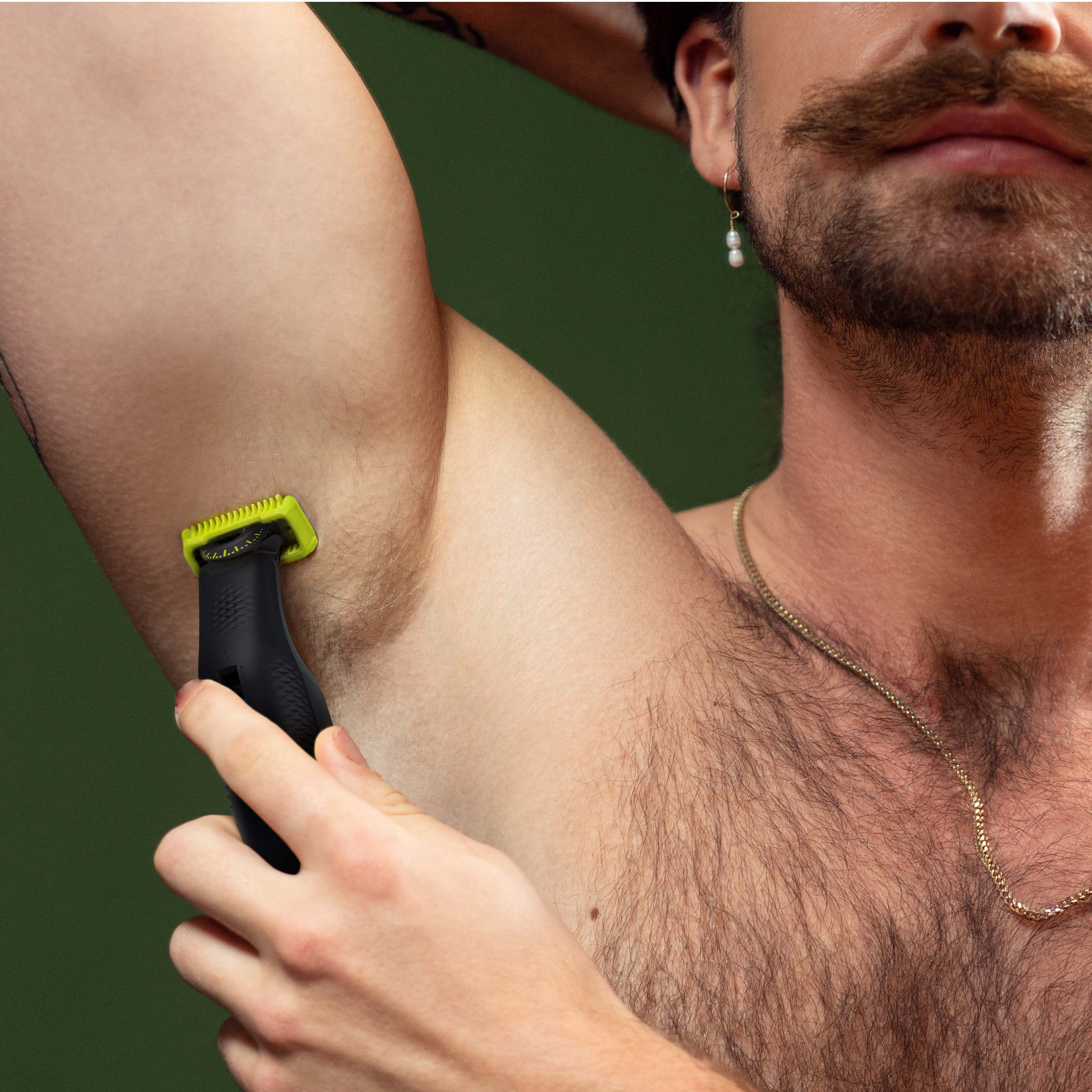 Mężczyzna goli pachę zieloną maszynką. Ciemnozielone tło.