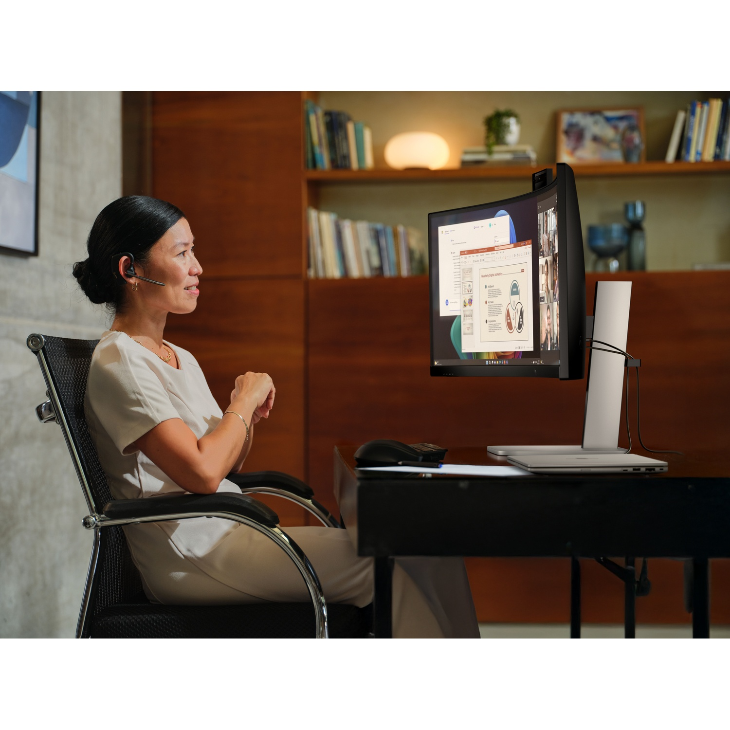 Vrouw zit aan bureau met headset, kijkt naar monitor met videoconferentie.