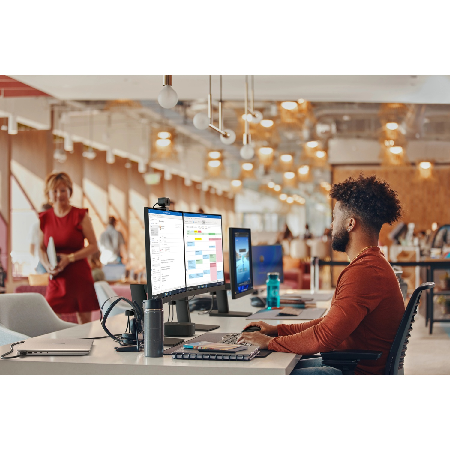 Homme à un bureau avec deux moniteurs, une femme en robe rouge passe au fond.