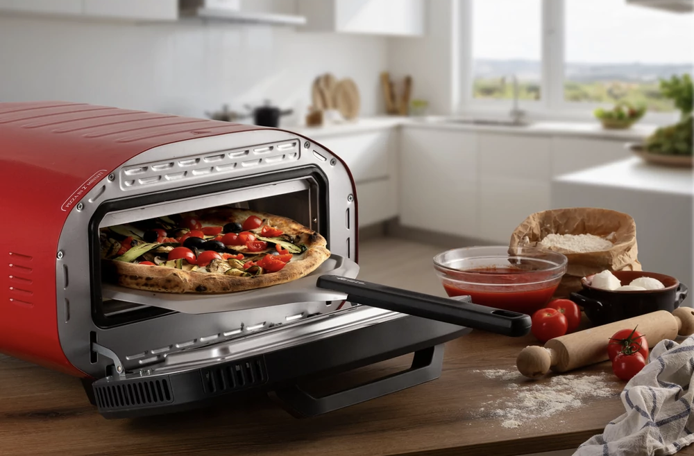 Egy piros sütő pizzával benne, mellette hozzávalók és konyhai eszközök.