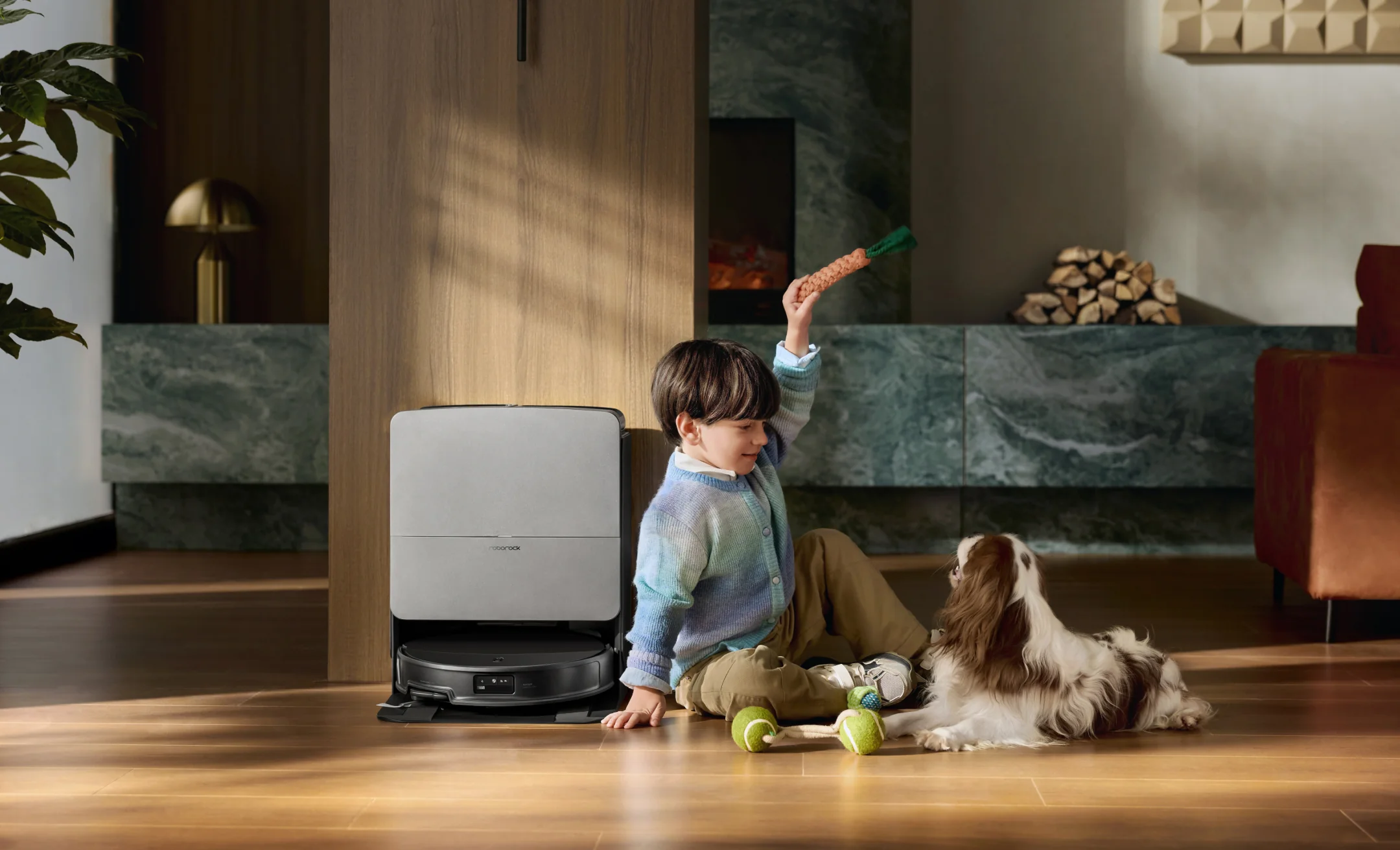 Un garçon joue avec un chien et un jouet devant un aspirateur robot.