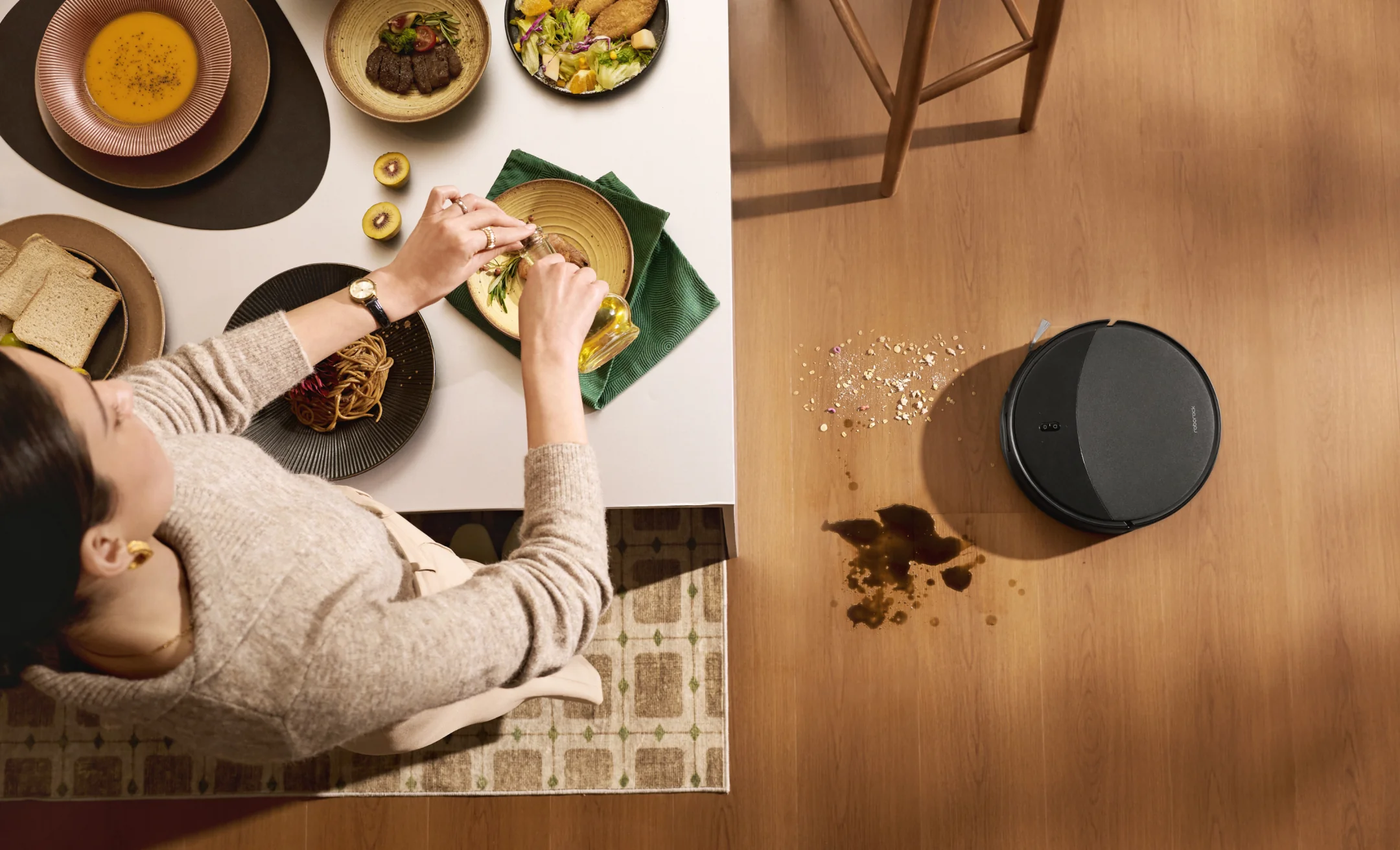 Un robot aspirateur nettoie de la nourriture renversée sur un parquet à côté d'une table avec de la nourriture.