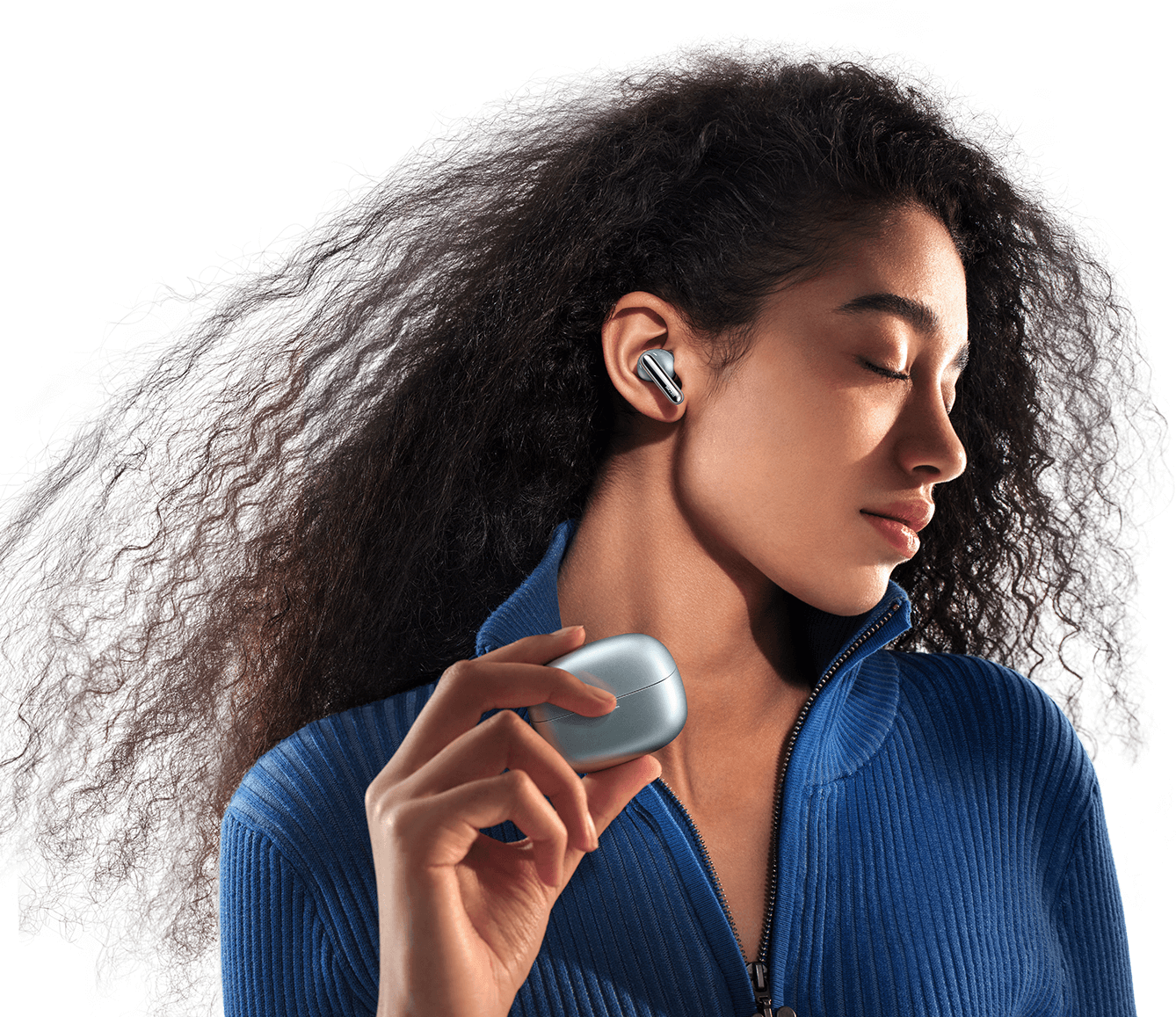 Una donna con capelli ricci indossa abiti blu e tiene in mano una custodia per auricolari argentata.