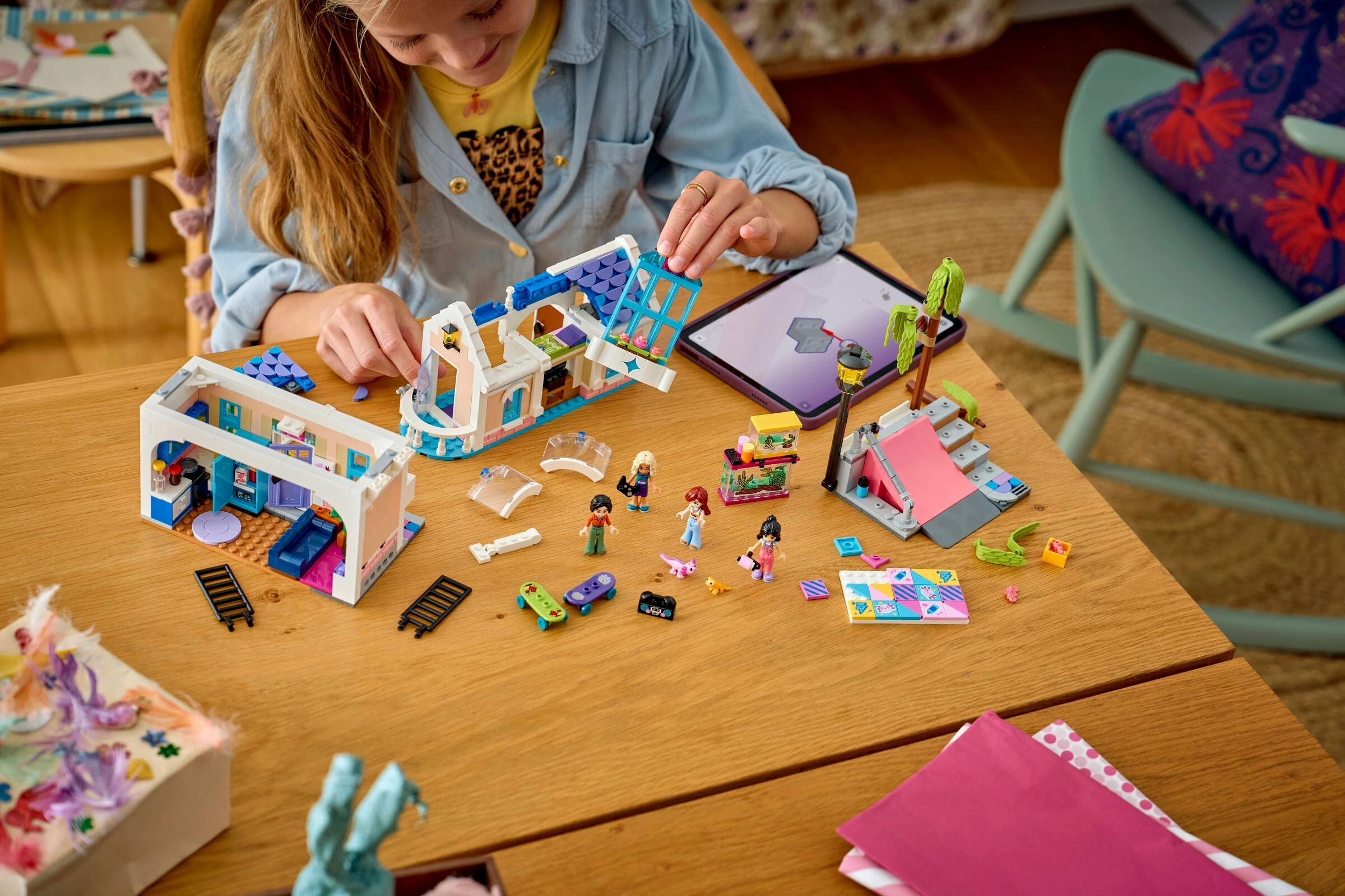 Dziewczynka buduje dom i rampę z klocków Lego z figurkami i deskorolkami na drewnianym stole.