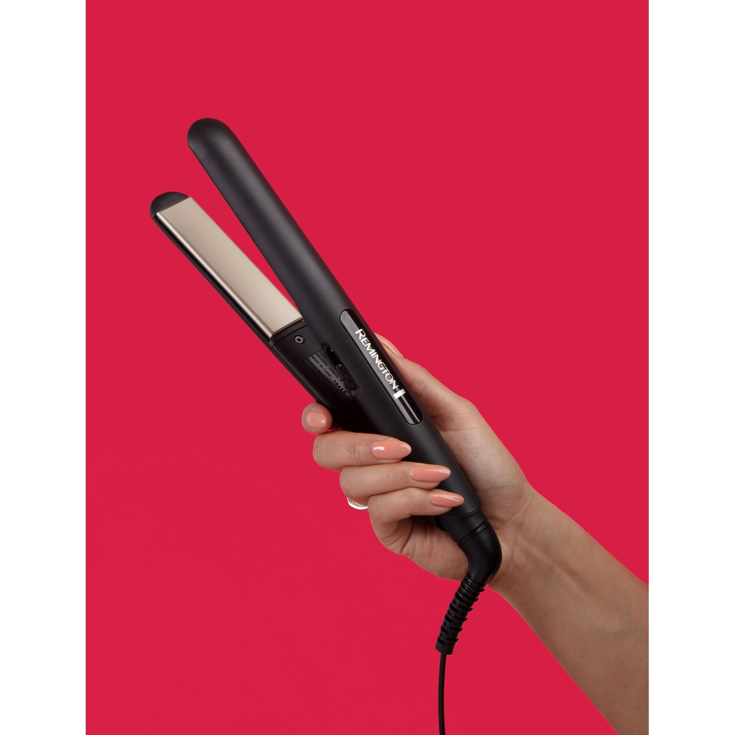 Una mano tiene una piastra per capelli Remington nera con piastre dorate su sfondo rosso.