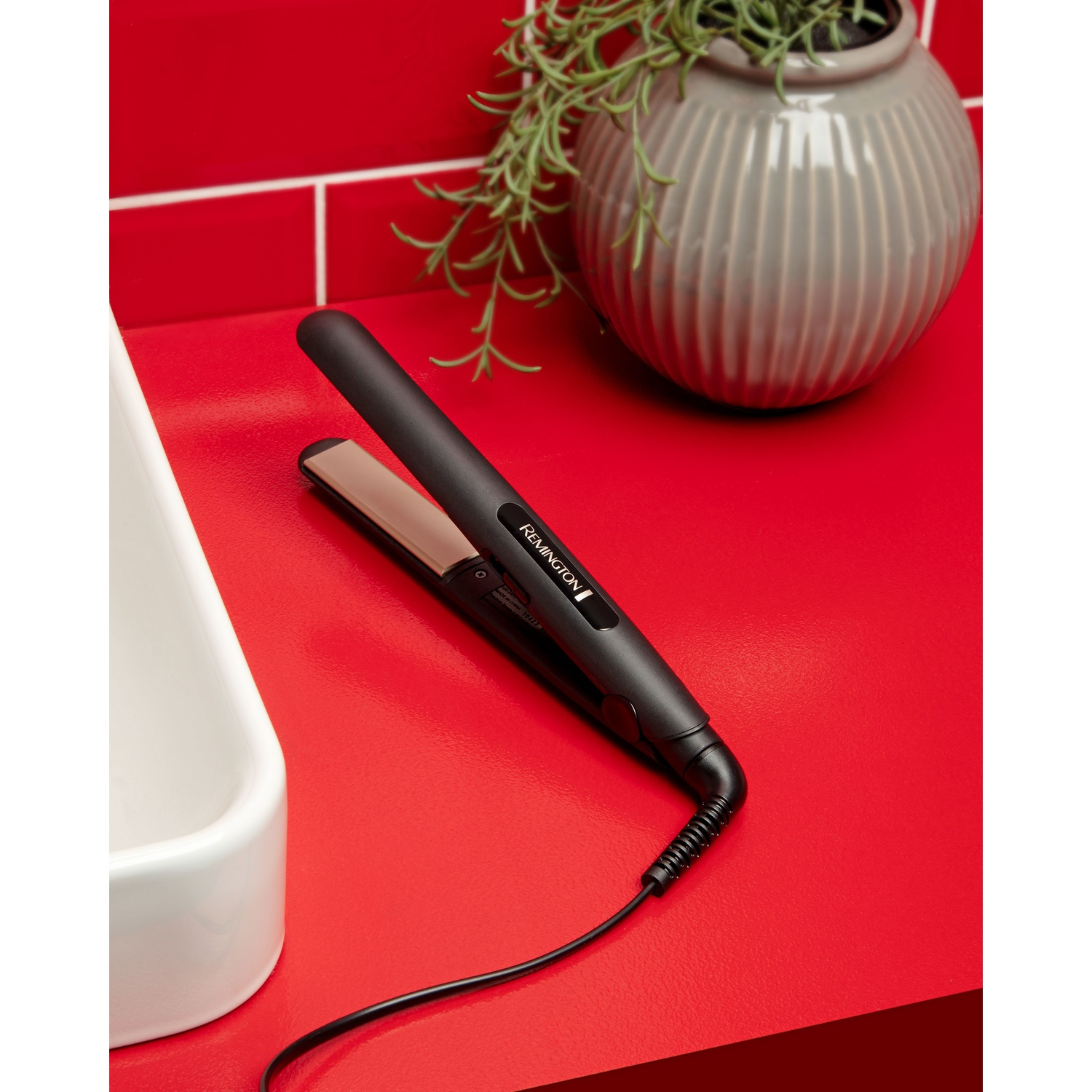 Una piastra per capelli Remington nera su superficie rossa, con vaso e pianta sullo sfondo.