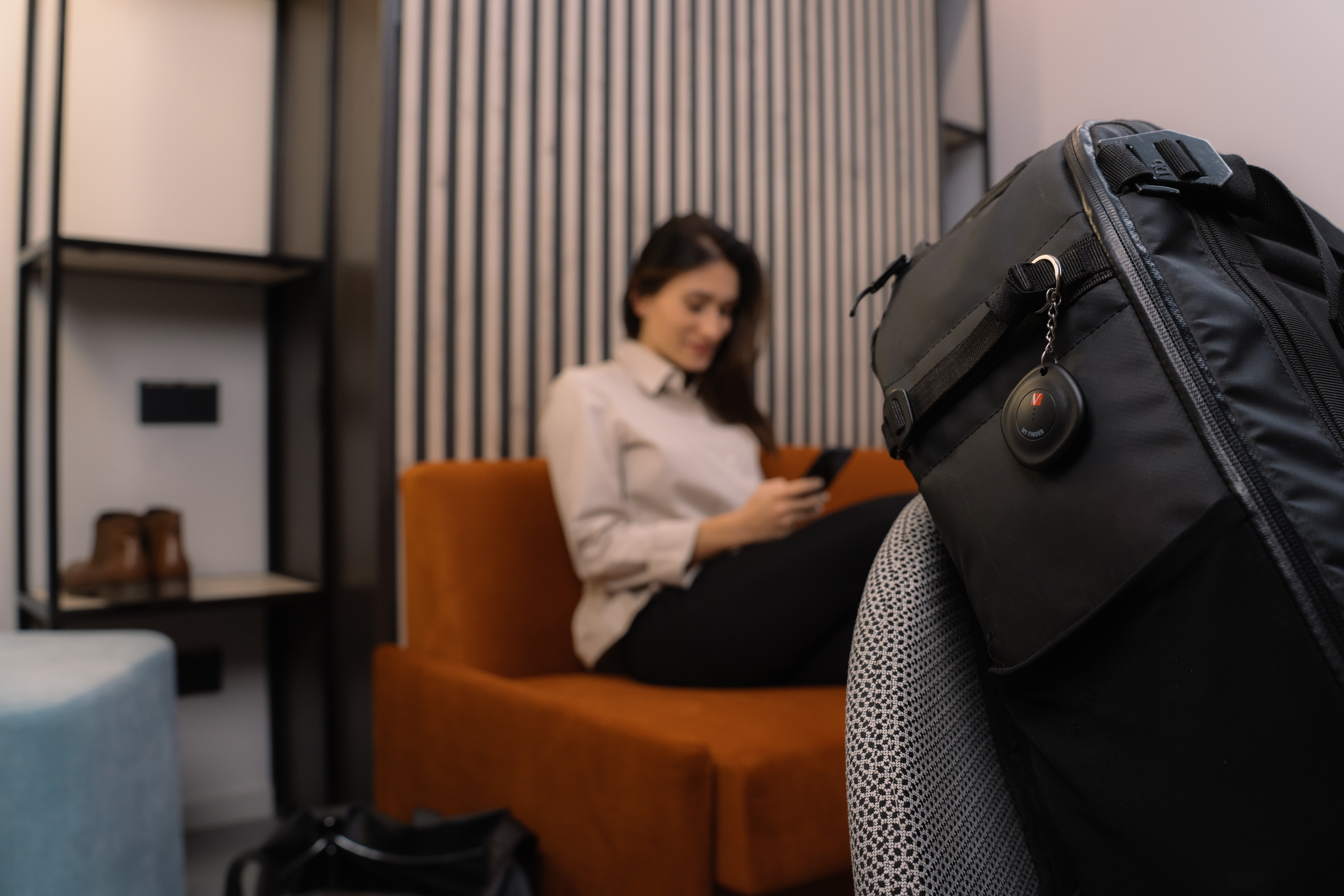 Eine Frau sitzt auf einem orangefarbenen Sessel und benutzt ihr Handy. Eine schwarze Tasche ist im Vordergrund.