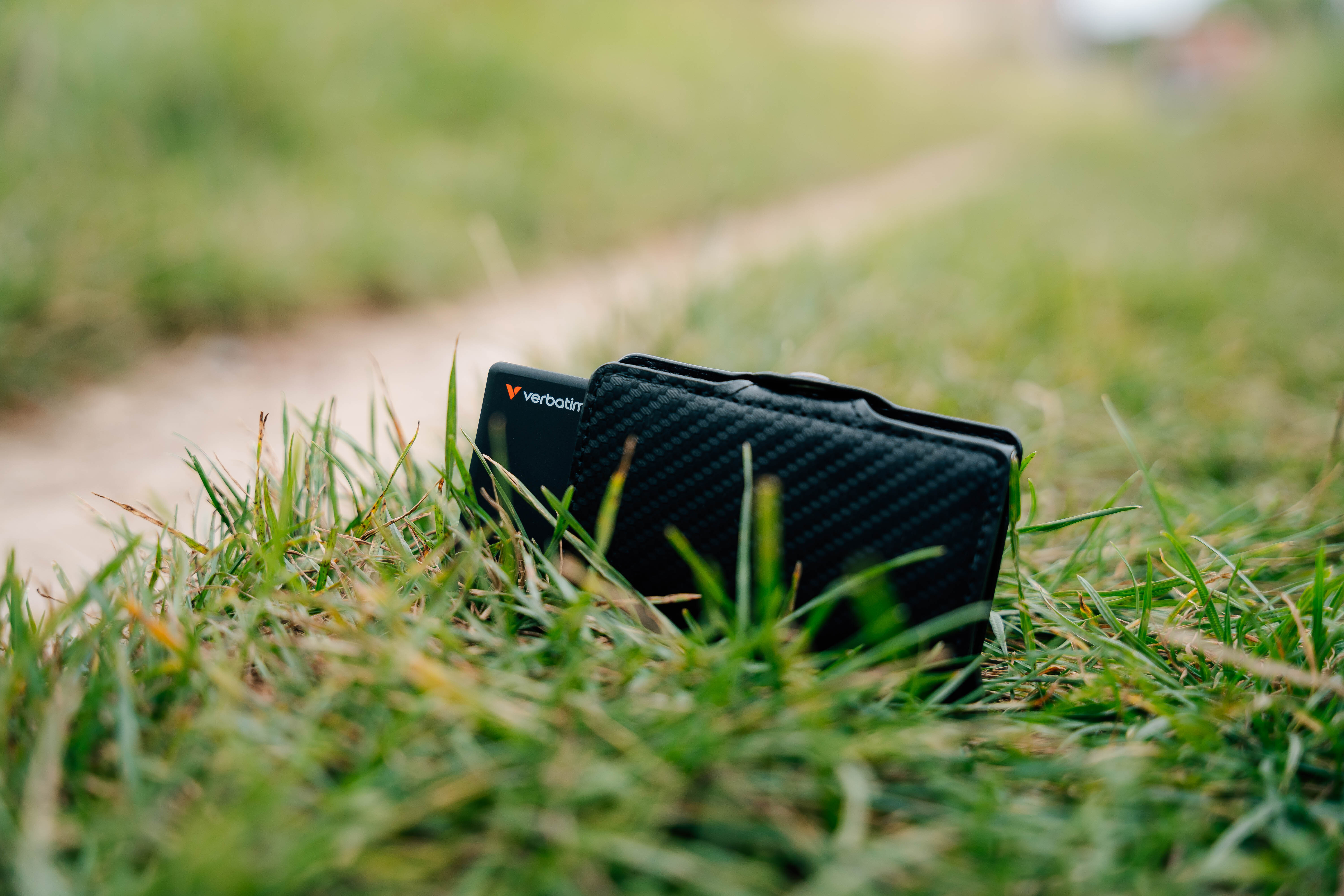 Ein schwarzes USB-Laufwerk mit Verbatim-Logo liegt im Gras neben einer Brieftasche.