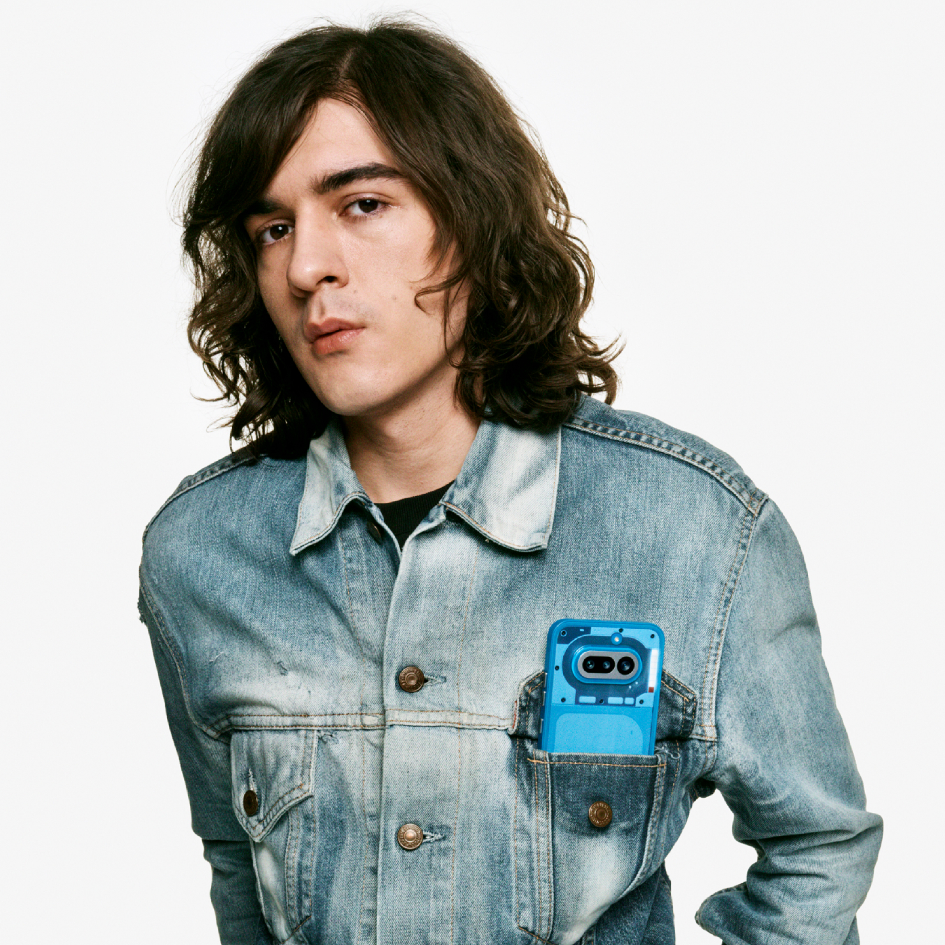 Un homme aux longs cheveux bruns porte une veste en jean avec un appareil bleu dans la poche poitrine.
