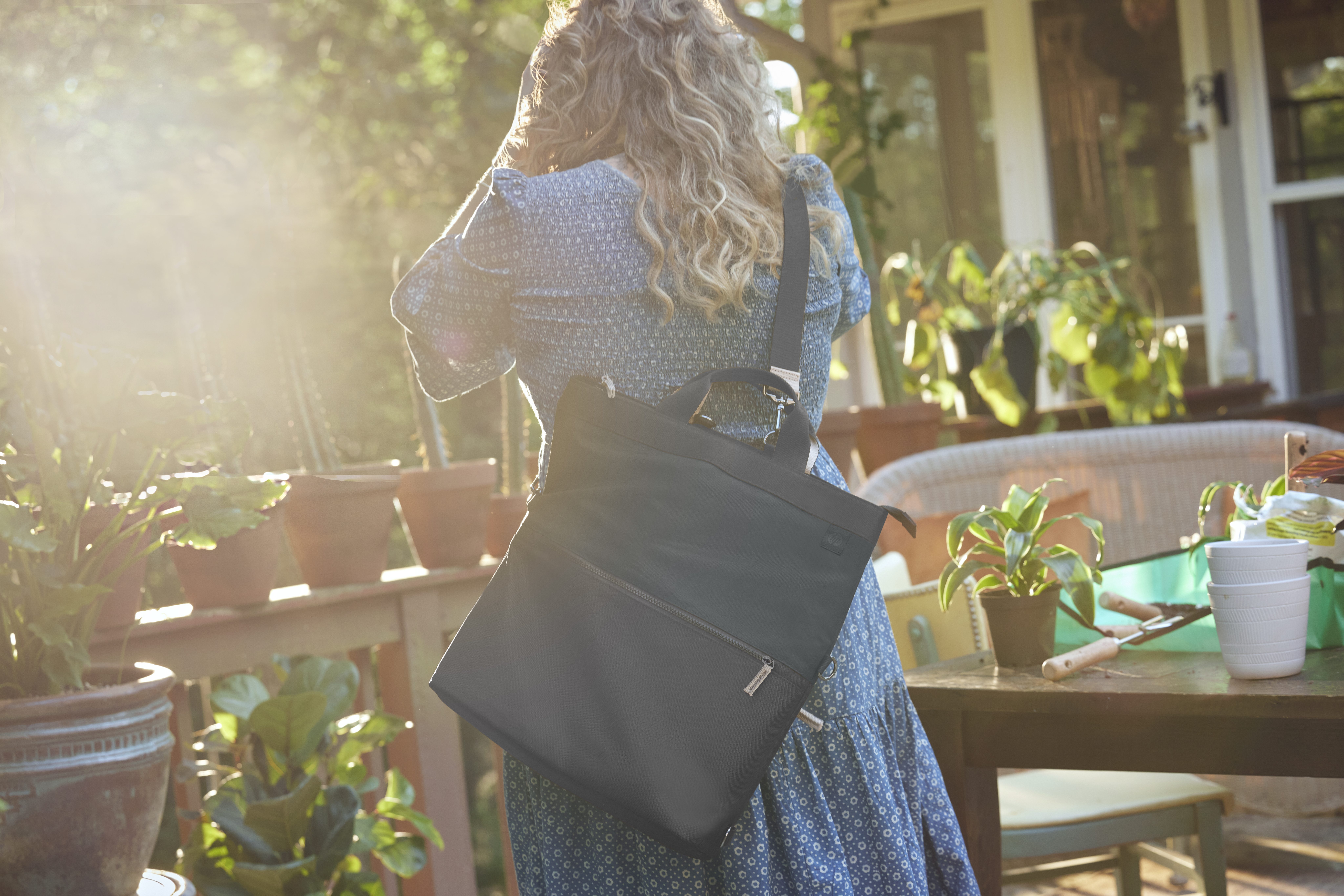 Mujer con pelo rizado en vestido azul y bolso negro en un porche con plantas.