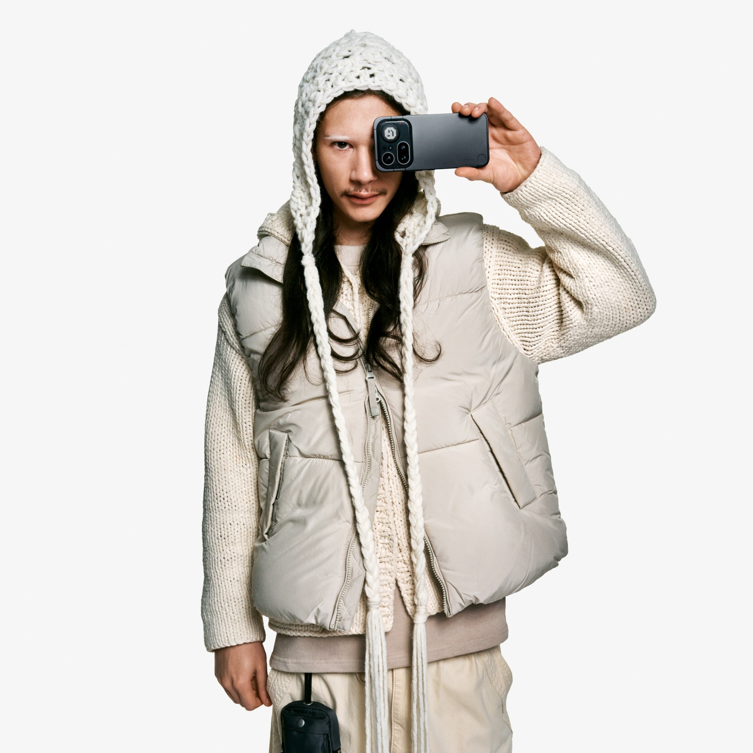 Persona con cappello di lana, gilet e maglione tiene uno smartphone davanti all'occhio.