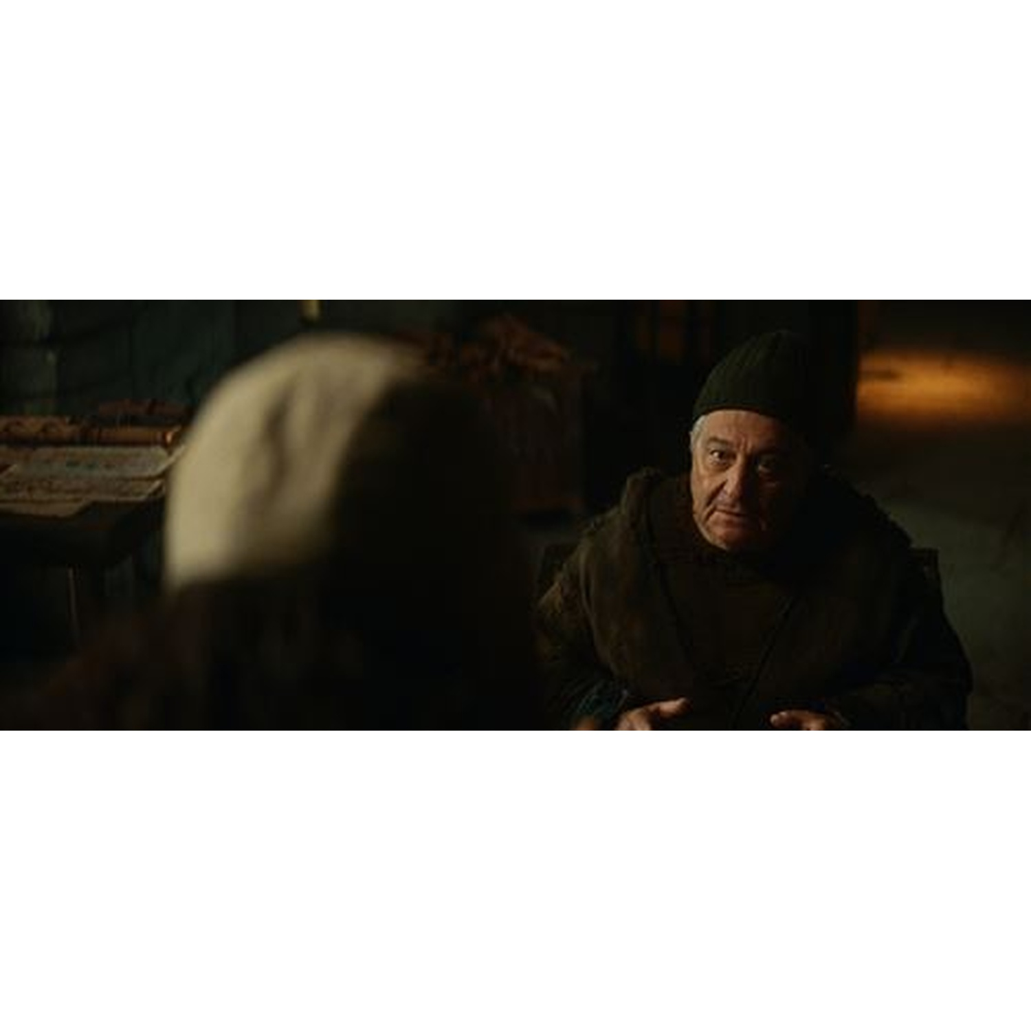 Un homme âgé en bonnet sombre et tunique marron parle à quelqu'un hors champ.