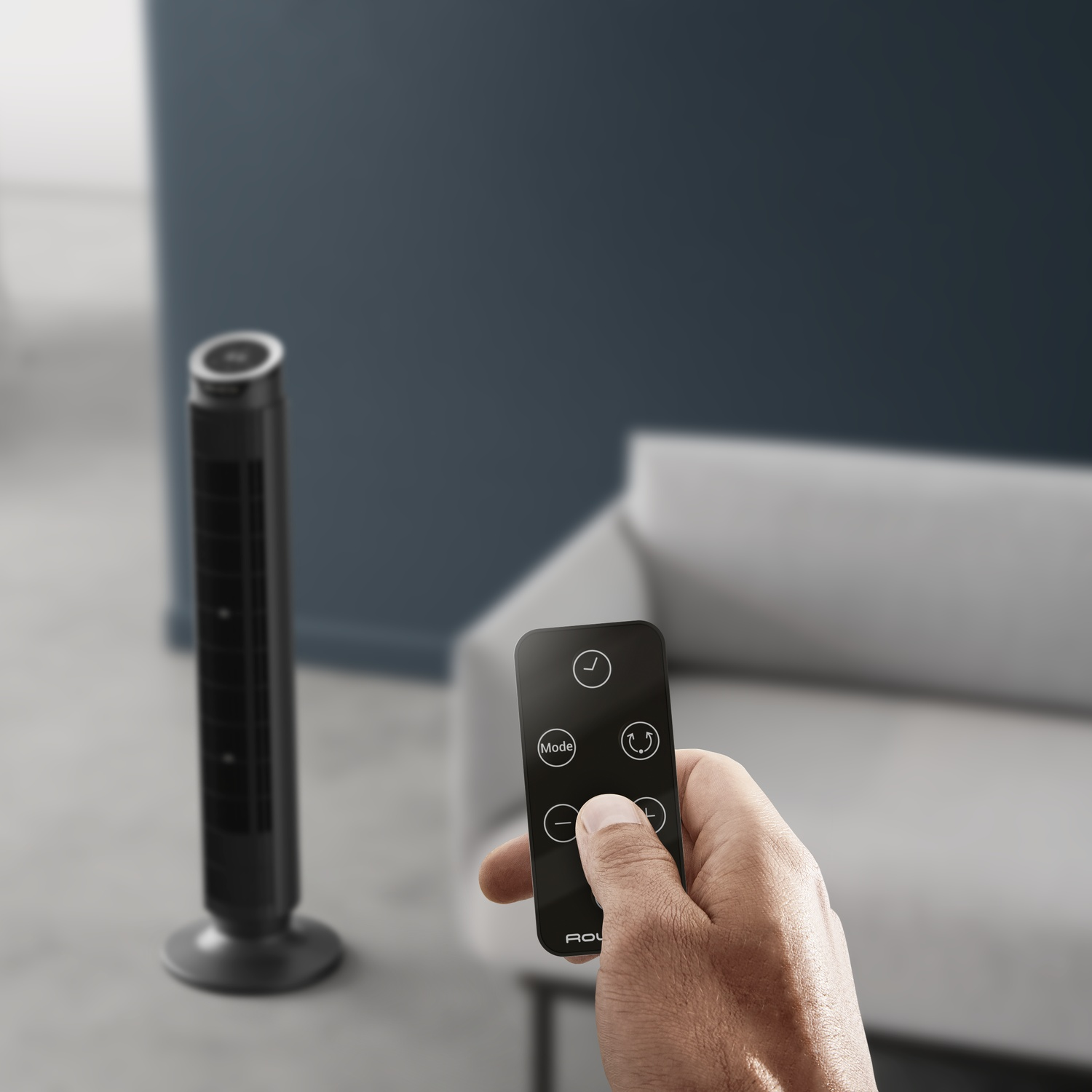 Une main tient une télécommande noire devant un ventilateur tour et un canapé.