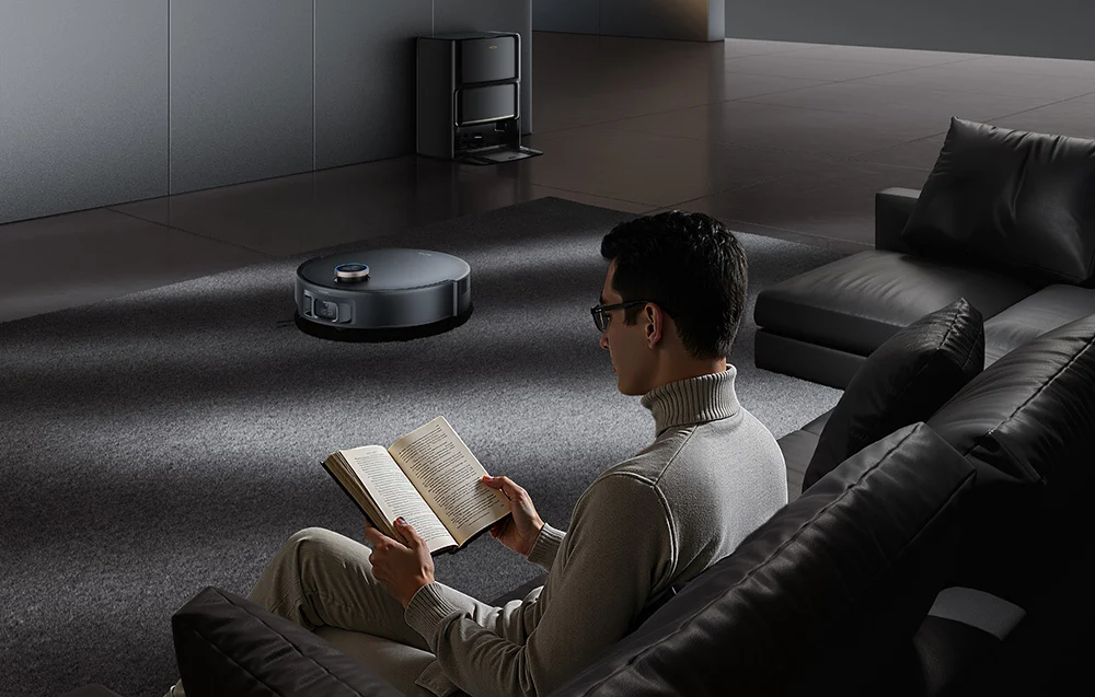 Egy férfi könyvet olvas, miközben egy robotporszívó takarít a háttérben.