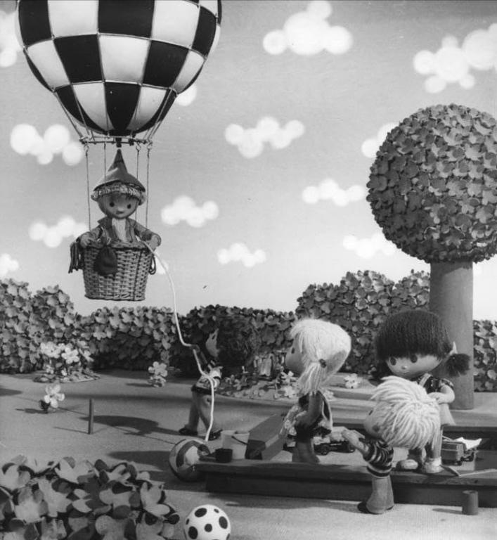 Eine Figur in einem Heißluftballon schwebt über Kindern, die auf einem Spielplatz spielen.
