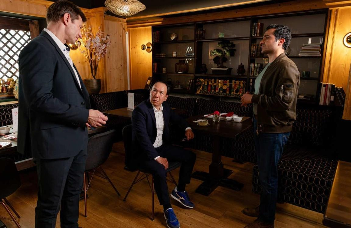 Drei Männer in einem Restaurant. Einer steht links, einer sitzt mittig, einer steht rechts.