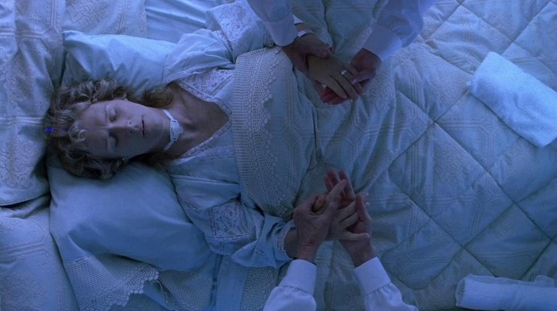 Eine Frau liegt im Bett, zwei Personen halten ihre Hände. Blaue Beleuchtung.