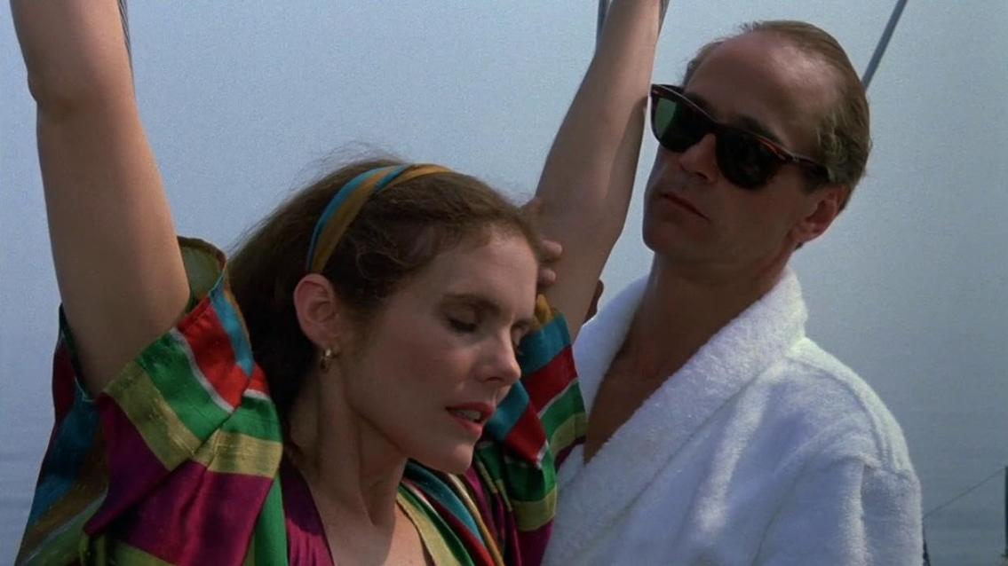Ein Mann mit Sonnenbrille und Bademantel hält eine Frau im bunten Kleid.