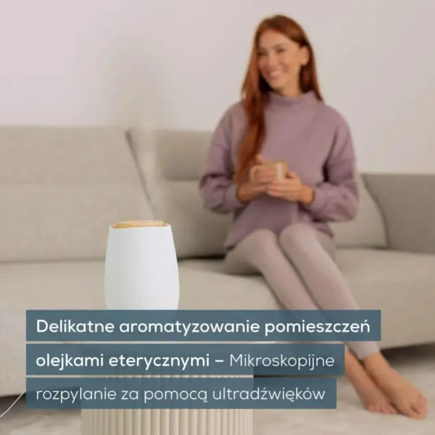 Kobieta siedzi na kanapie z kubkiem, w tle dyfuzor zapachowy.