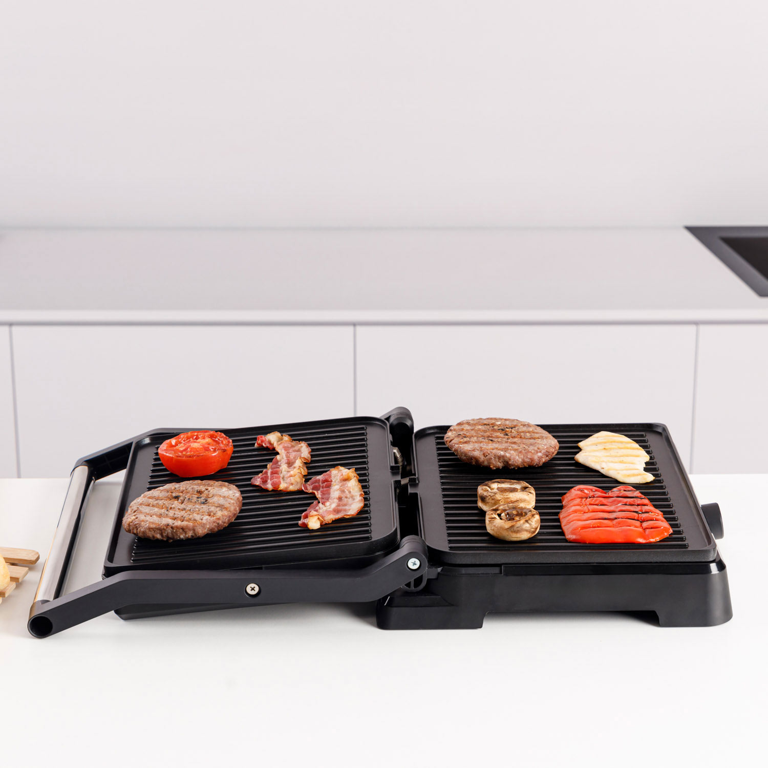 Una parrilla eléctrica abierta con hamburguesas, tocino, tomates, champiñones y pimientos.