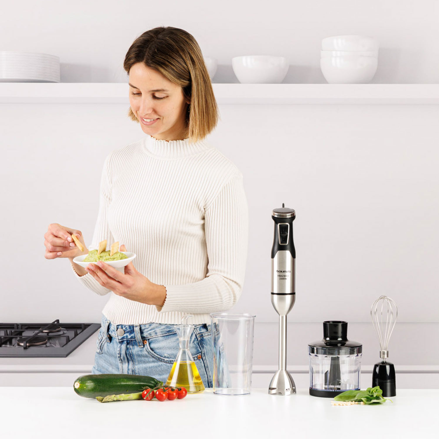Una mujer con suéter blanco sostiene un cuenco de dip y patatas. Utensilios de cocina en la encimera.