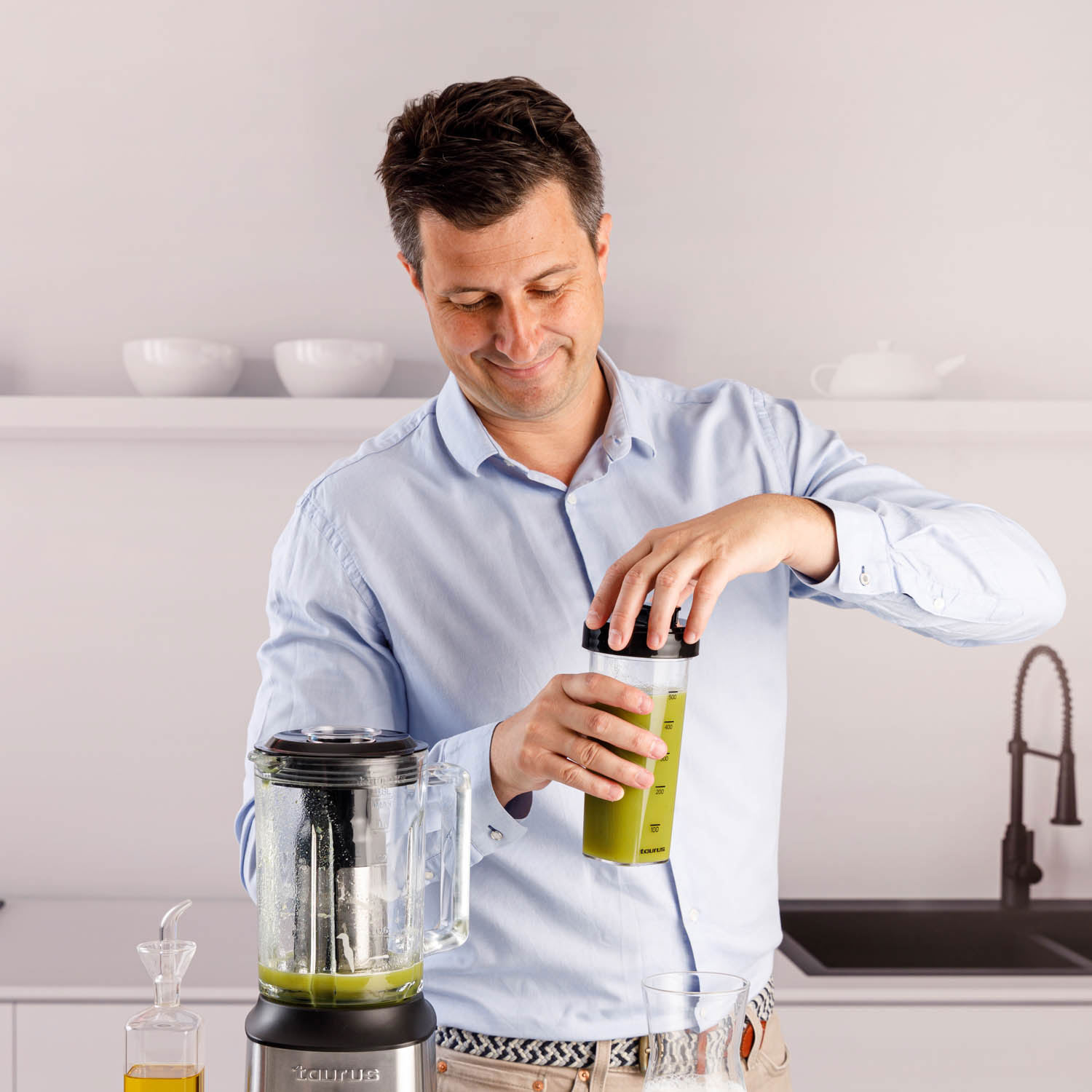 Hombre con camisa azul cierra envase con jugo verde junto a licuadora.