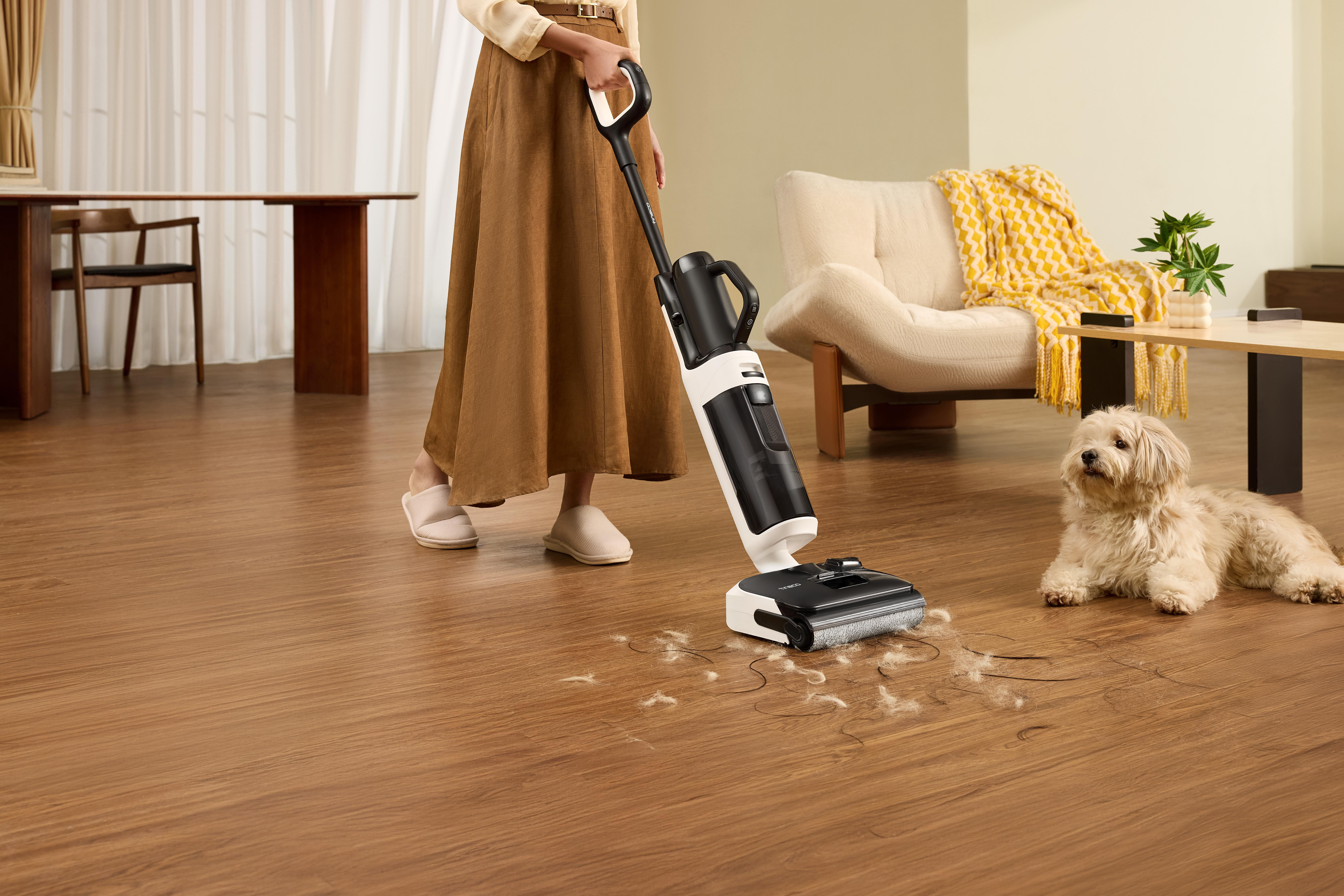 Eine Person saugt mit einem kabellosen Staubsauger auf einem Holzboden. Ein Hund liegt daneben.