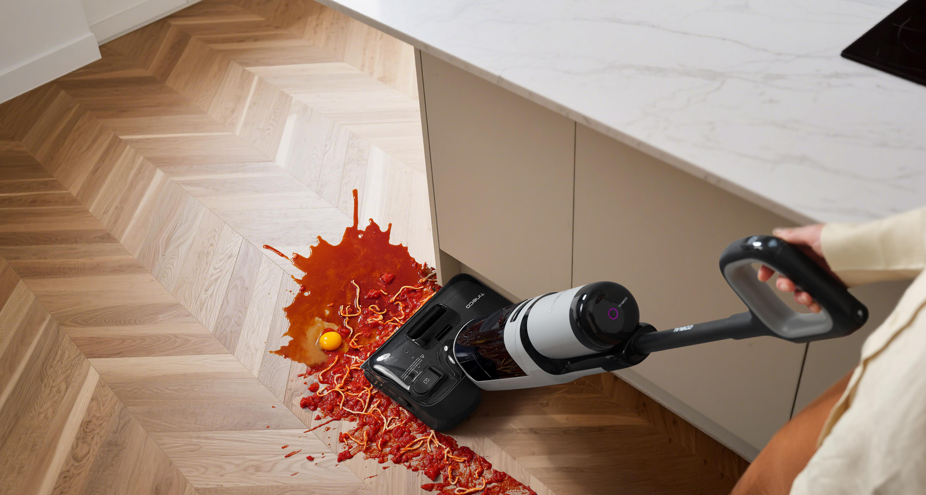 Ein Staubsauger reinigt verschüttete Spaghetti, Tomatensoße und ein Ei auf einem Holzboden.