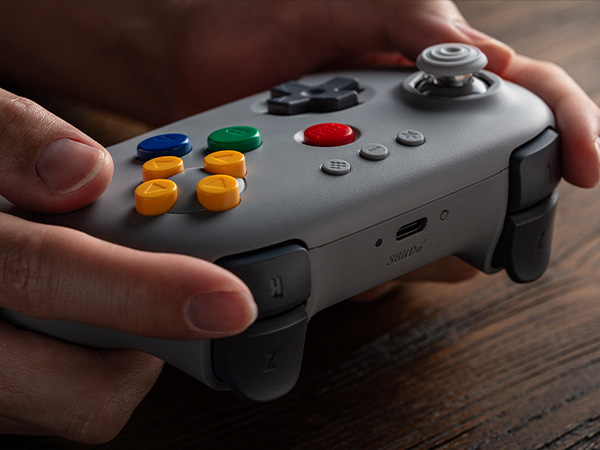 Grauer Gamecontroller in Händen, mit bunten Knöpfen und Joysticks.