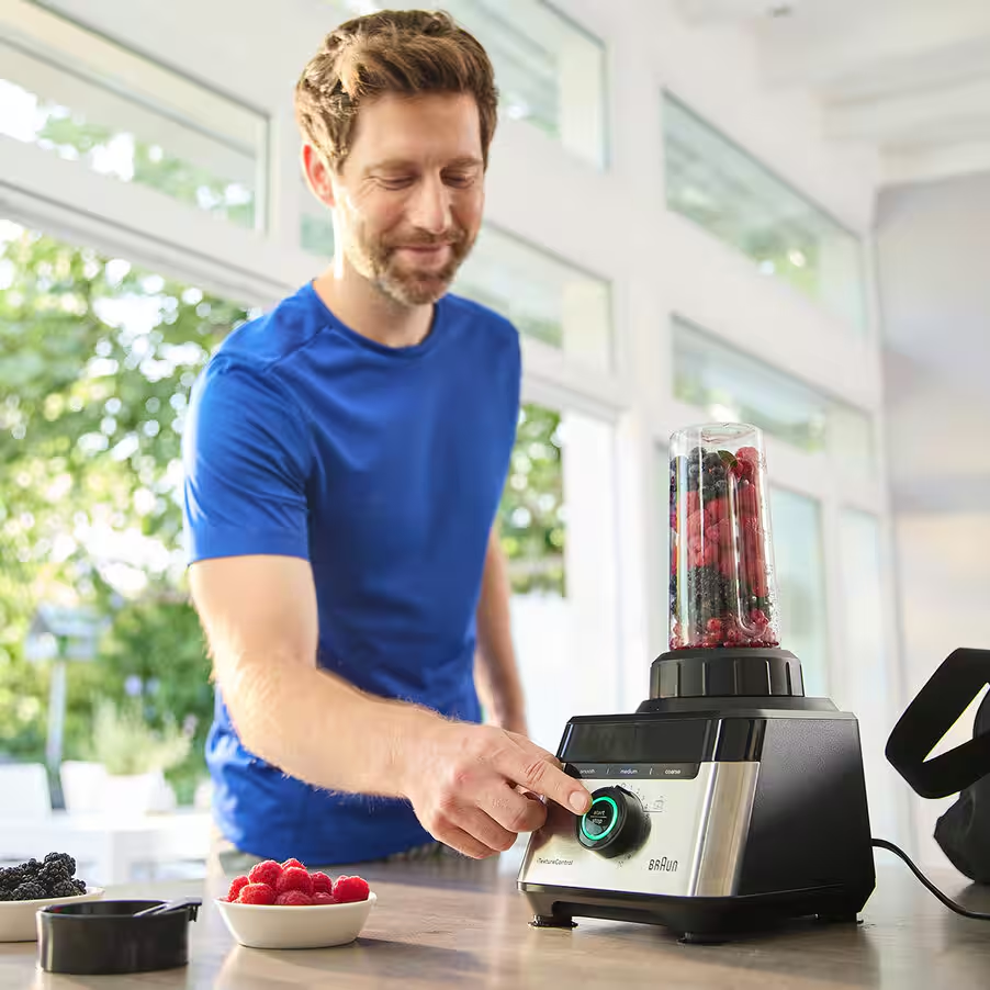 Mann in blauem T-Shirt bedient Mixer mit Beeren. Schale mit Himbeeren.