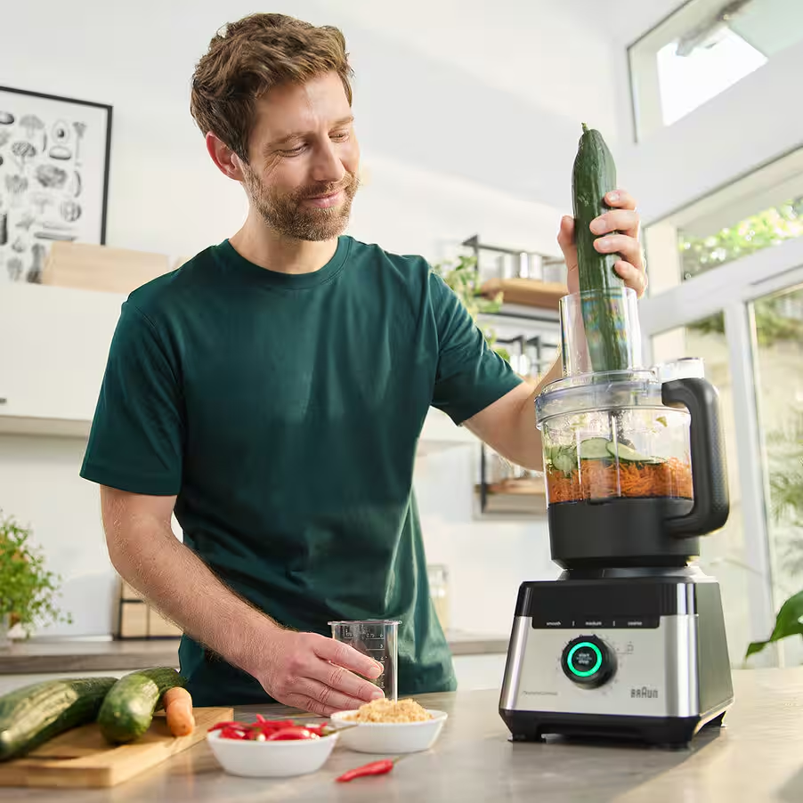 Mann gibt Gurke in Küchenmaschine mit Karotten und Chilis auf der Arbeitsplatte.
