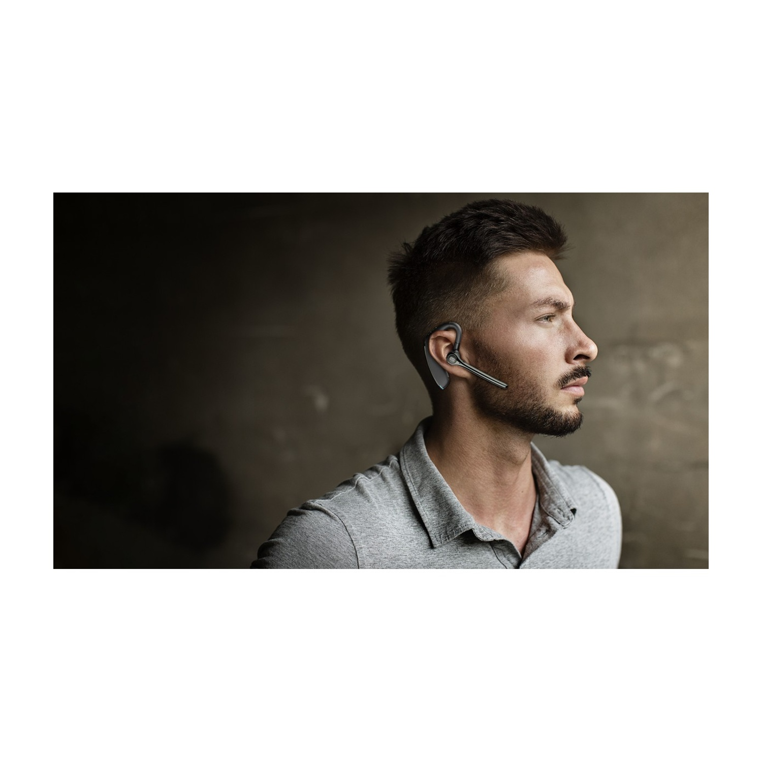 Uomo con auricolare Bluetooth nero all'orecchio sinistro, polo grigia, sfondo scuro.