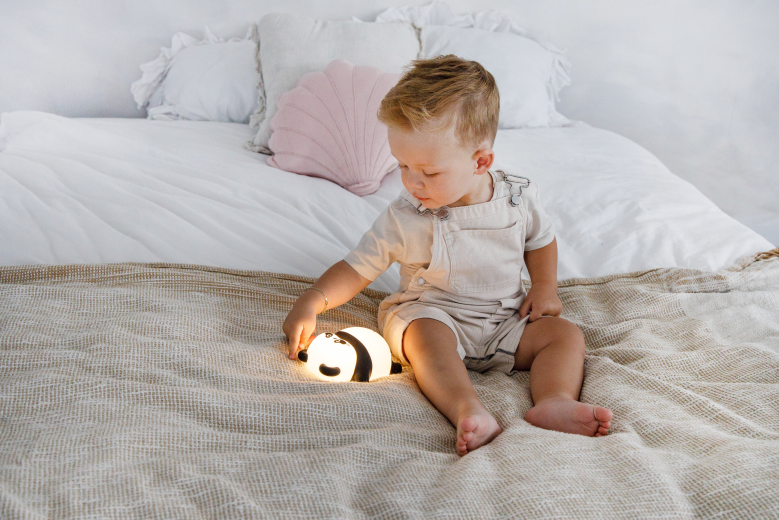 Maluch w ogrodniczkach siedzi na łóżku, bawiąc się świecącą lampką nocną panda.