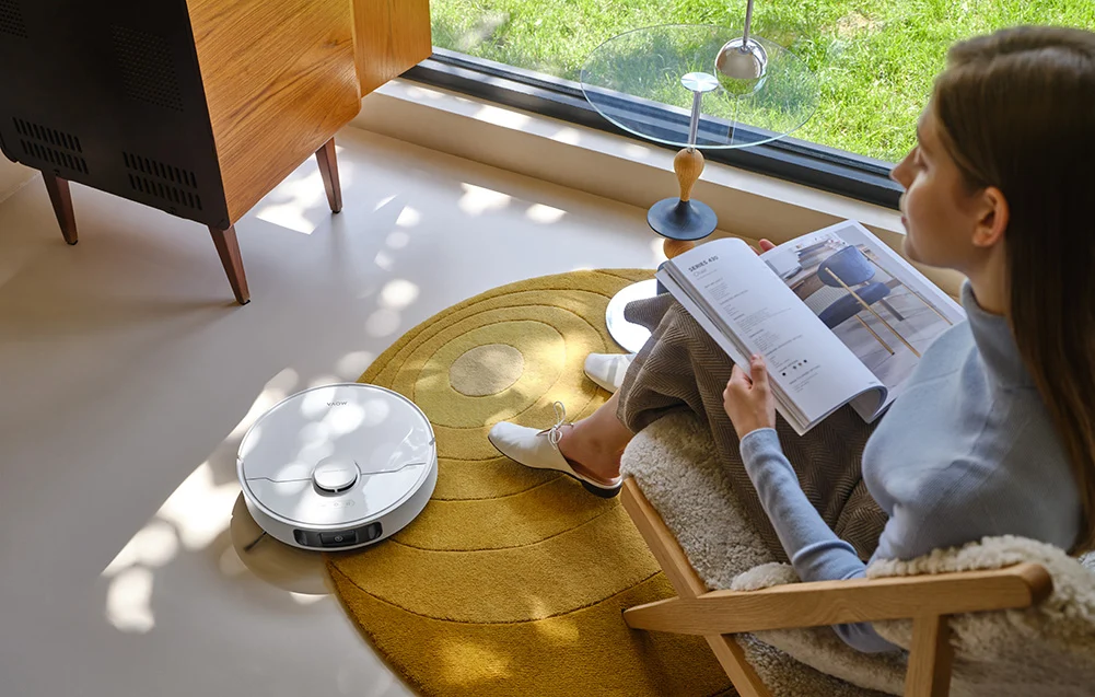 Egy fehér robotporszívó sárga szőnyegen, fa szekrény mellett.