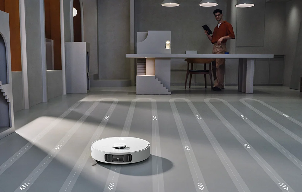 Egy fehér robotporszívó takarít egy modern szobában, háttérben egy férfival.
