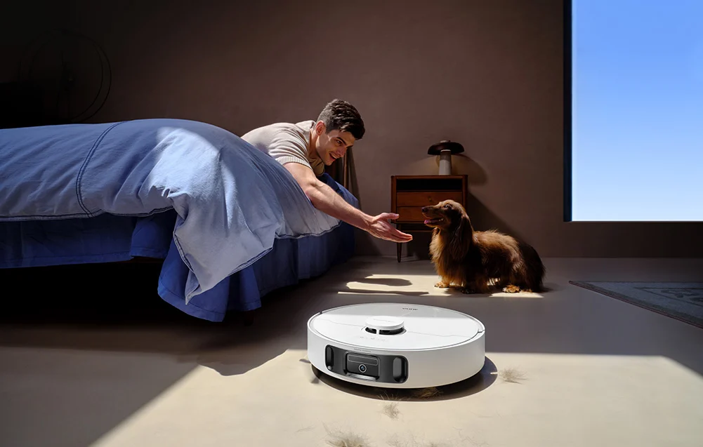 Férfi nyújtja a kezét kutyának robotporszívó és ágy mellett.