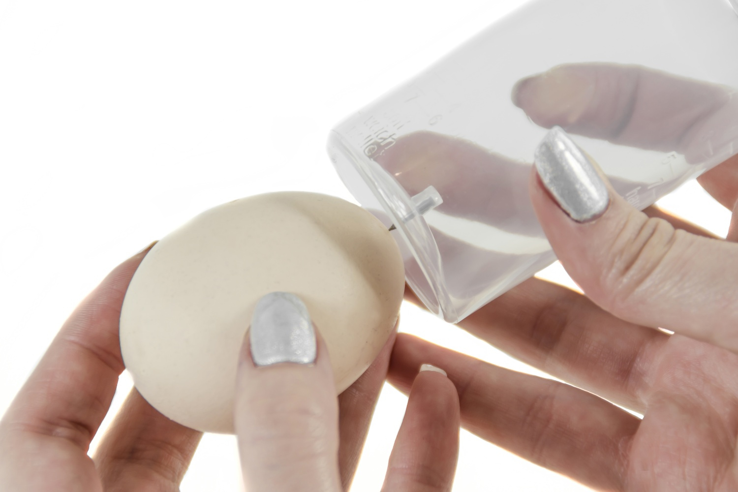 Dłoń z srebrnymi paznokciami trzyma jasno brązowe jajko i przezroczysty plastikowy pojemnik.