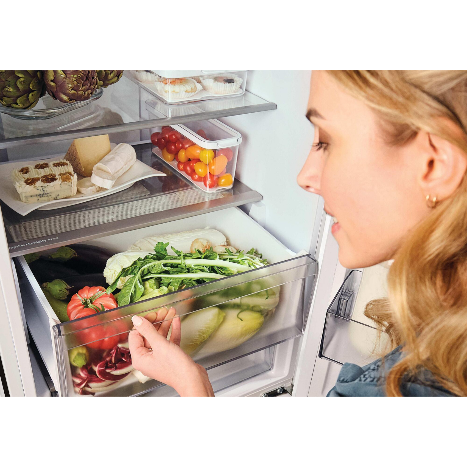 Una mujer abre un cajón de refrigerador con verduras. Se ven queso y alcachofas.