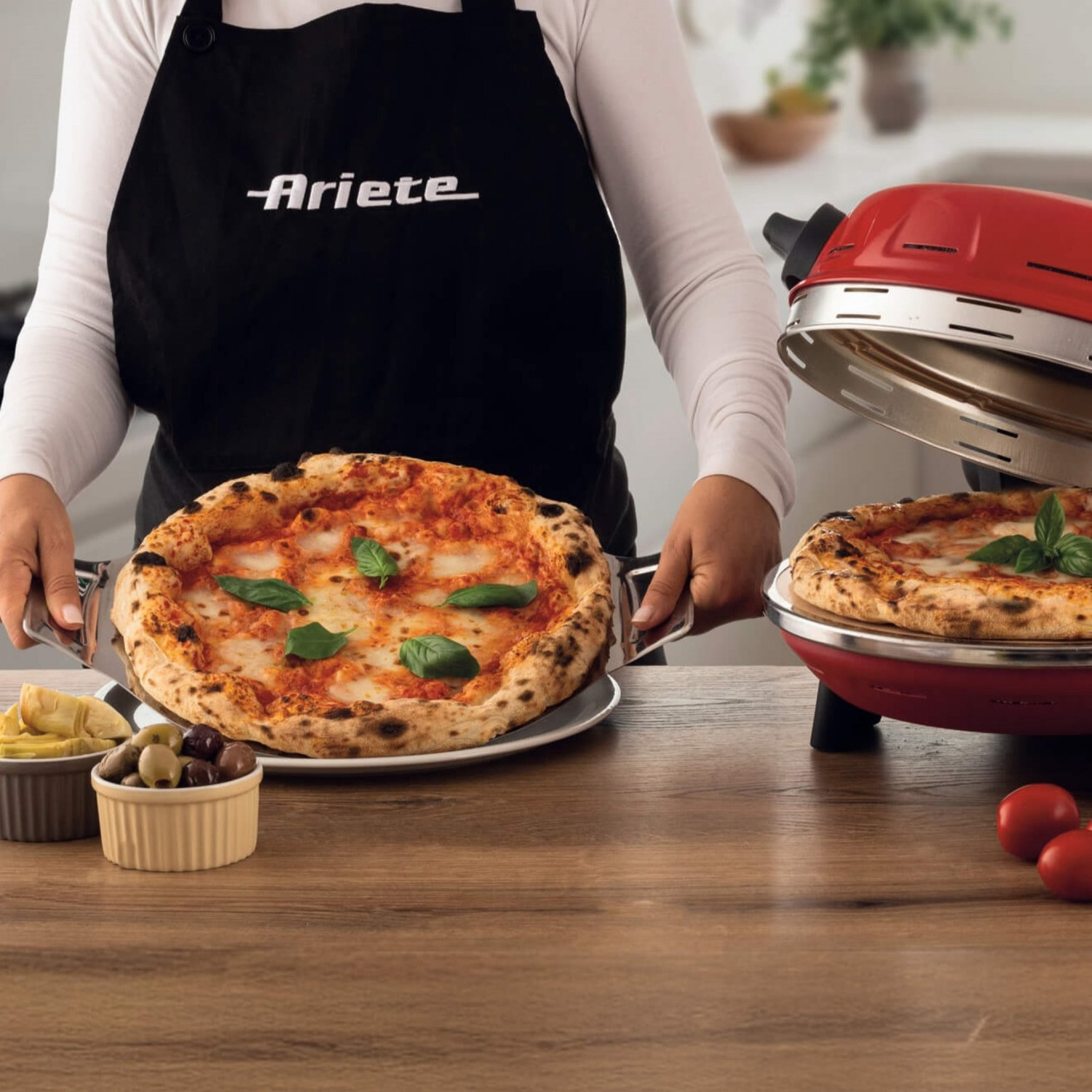 Una persona con grembiule nero presenta due pizze accanto a un forno per pizza rosso.