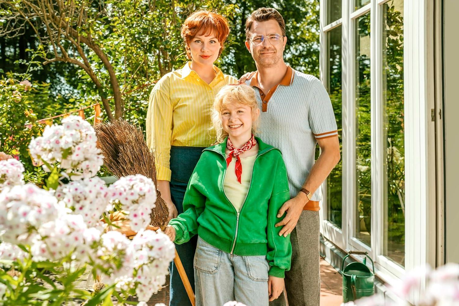 Eine Familie posiert im Garten vor weißen Blumen. Die Frau trägt ein gelbes Hemd, das Kind einen grünen Hoodie.