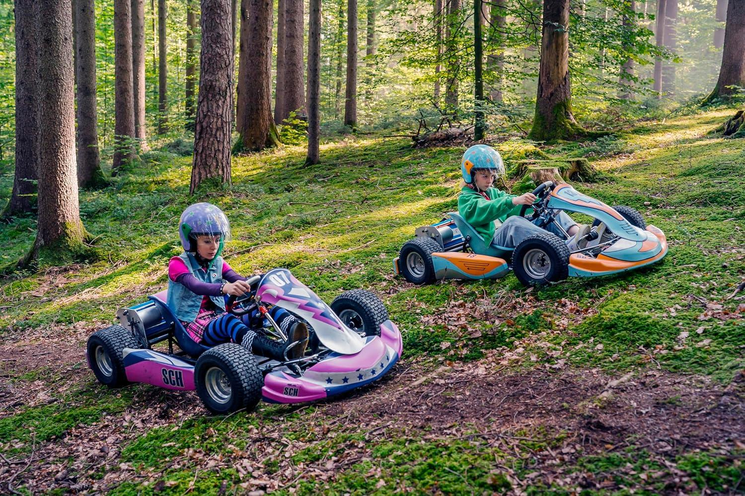 Zwei Kinder in Helmen rasen mit Go-Karts durch einen sonnigen Wald mit moosigem Boden.