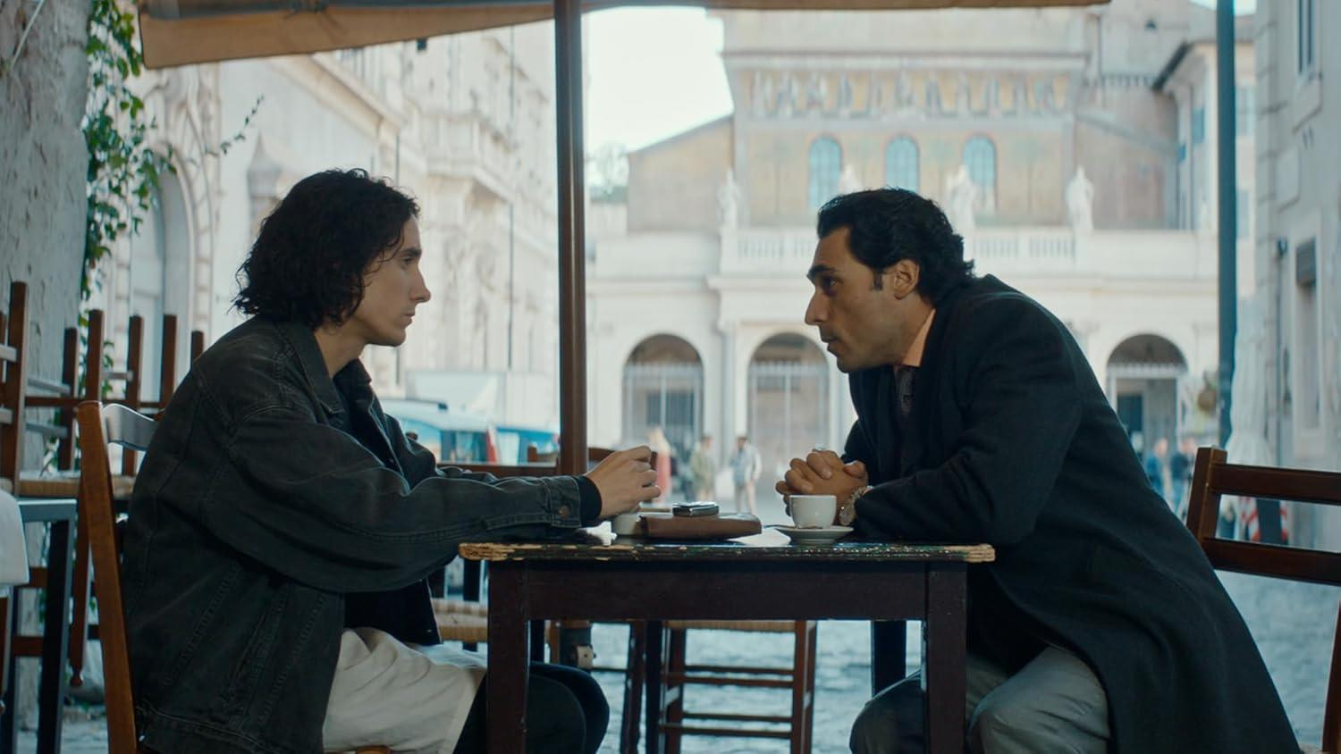 Zwei Männer sitzen an einem Tisch im Freien und trinken Kaffee. Im Hintergrund ist eine Stadtansicht.